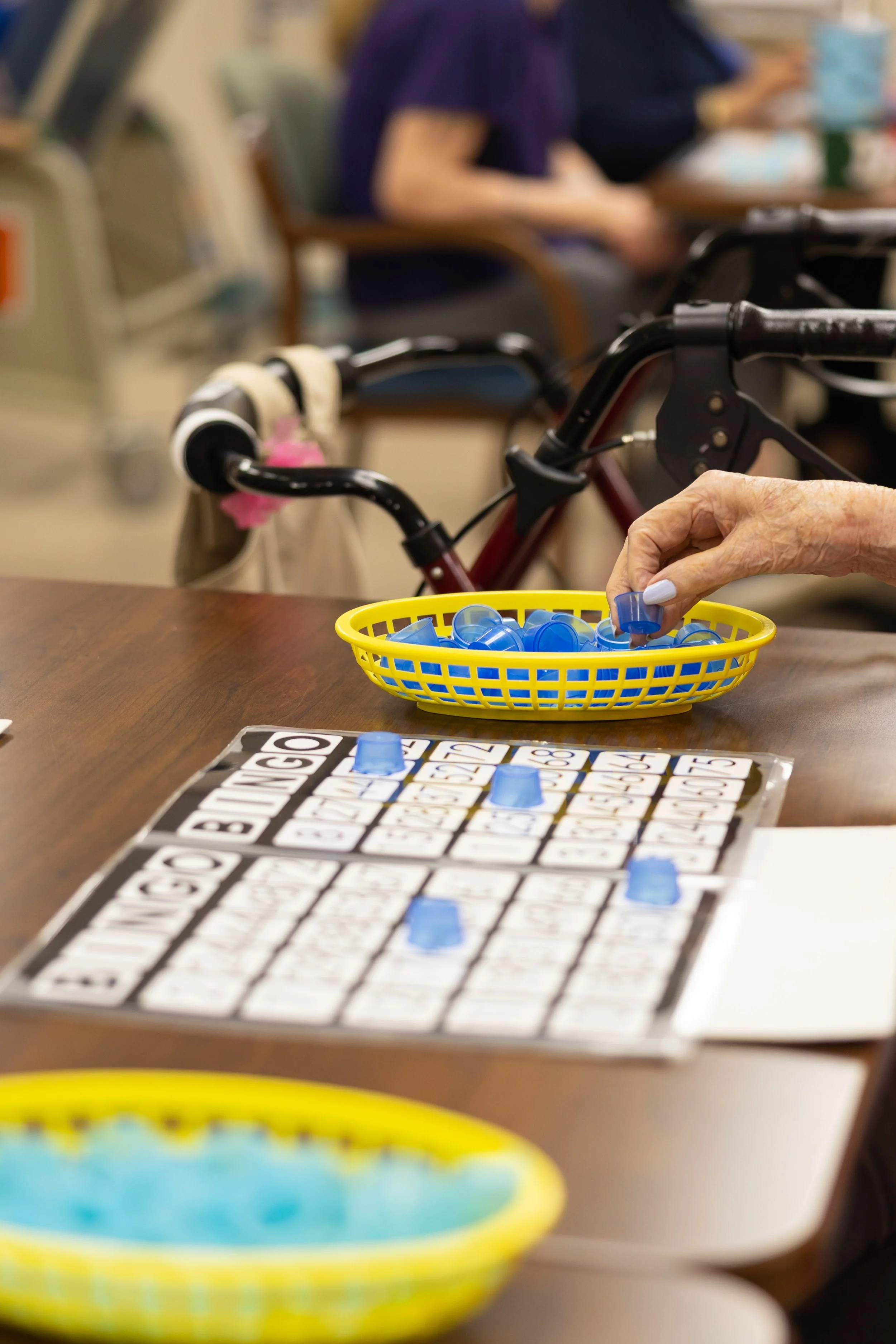 Elderly person playing bingo in a nursing home with blue plastic bingo chips and a game card on a wooden table.
