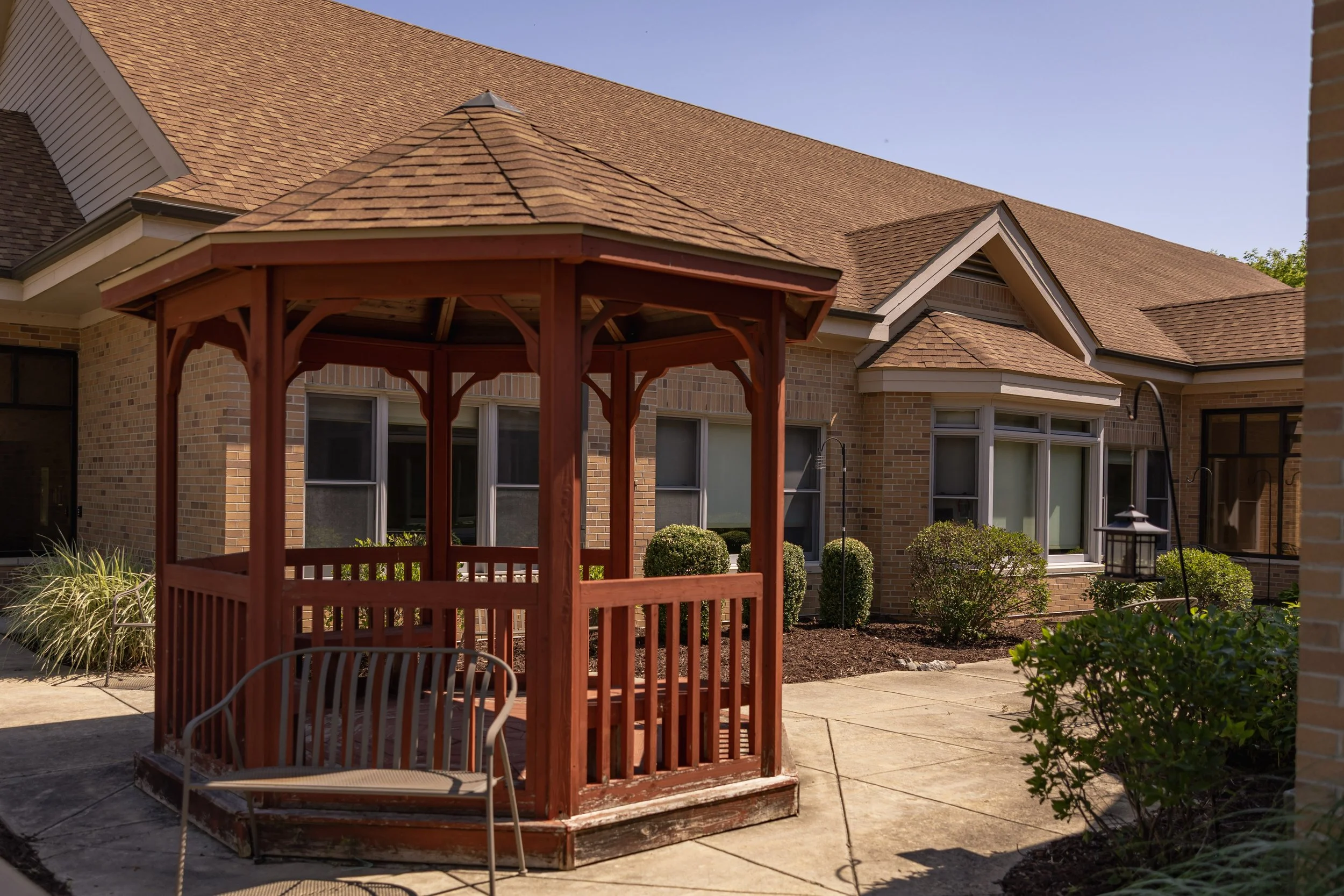 A red wooden gazebo with a bench in front, located in the backyard of a brick house with multiple windows and landscaping shrubs.
