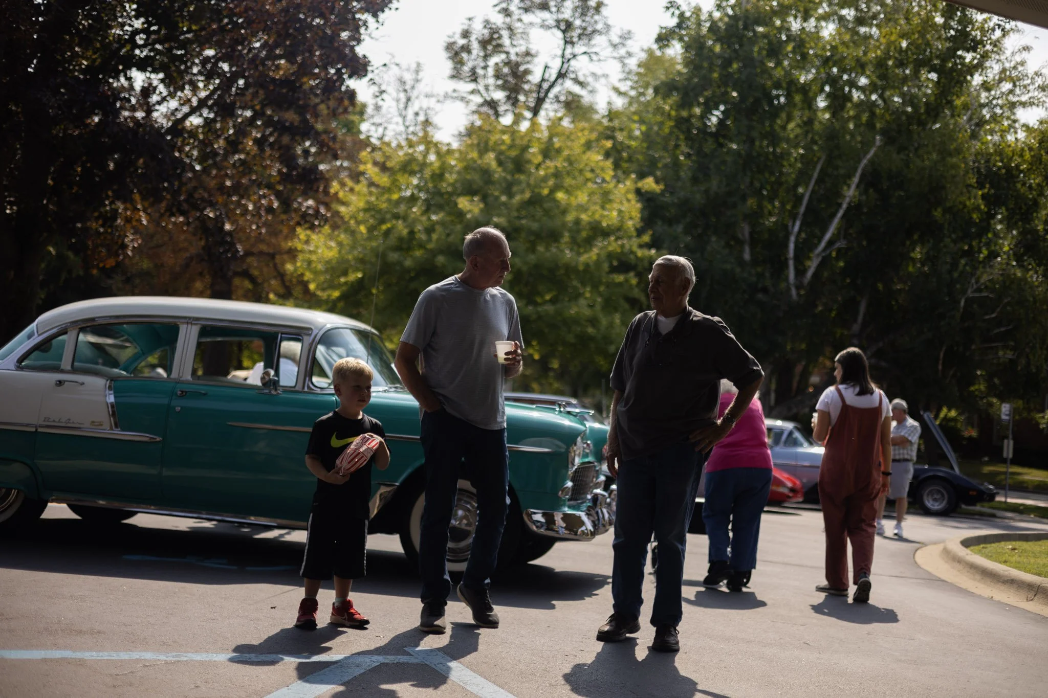 A group of people is gathered in a parking lot outdoors, with vintage cars and trees in the background. The sunlight creates silhouettes of the individuals, including two men talking, a boy holding a snack, and other people walking around.