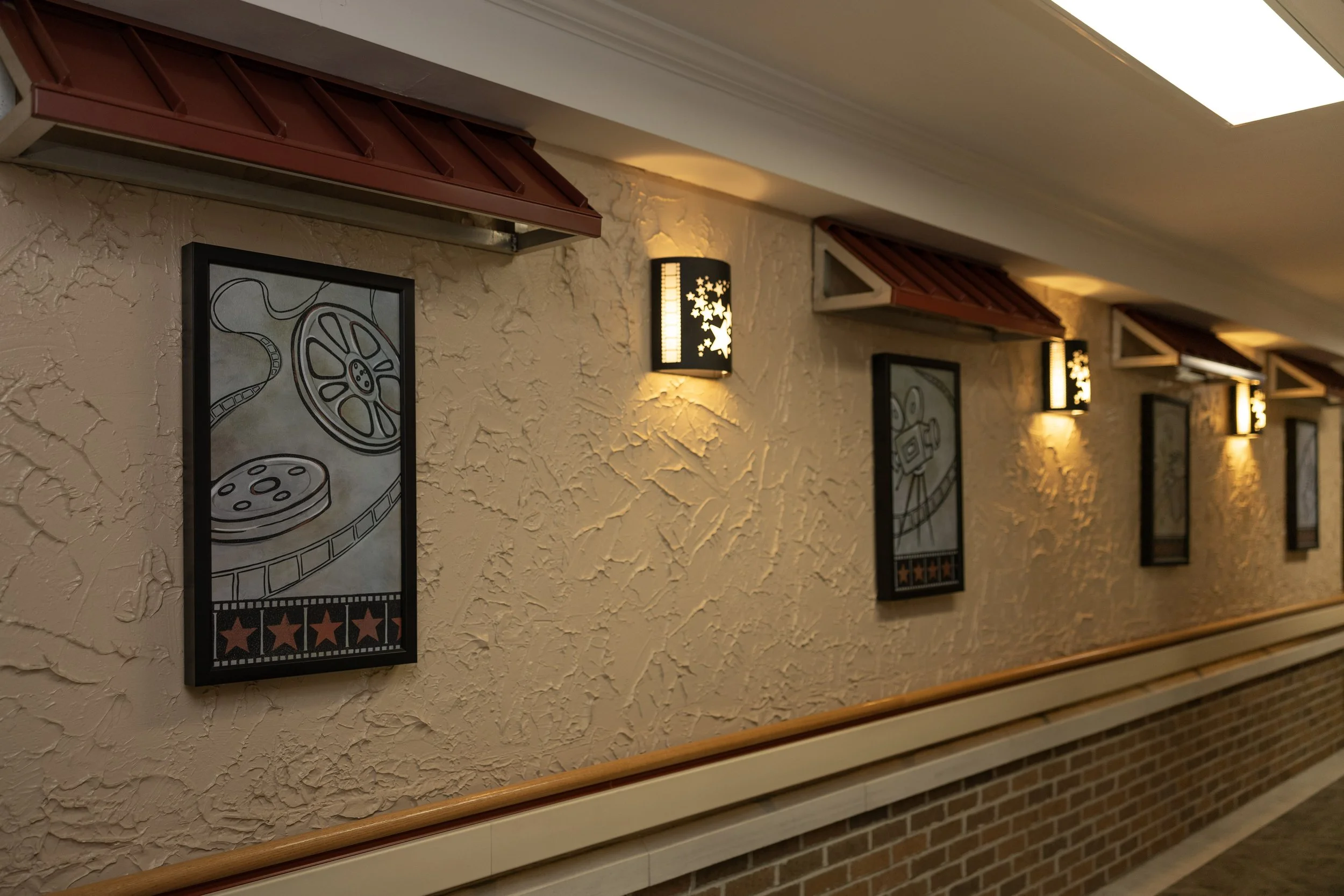 Decorative movie-themed wall art with film reels, projectors, and stars, illuminated by wall sconces in a hallway with textured walls and brick baseboard.
