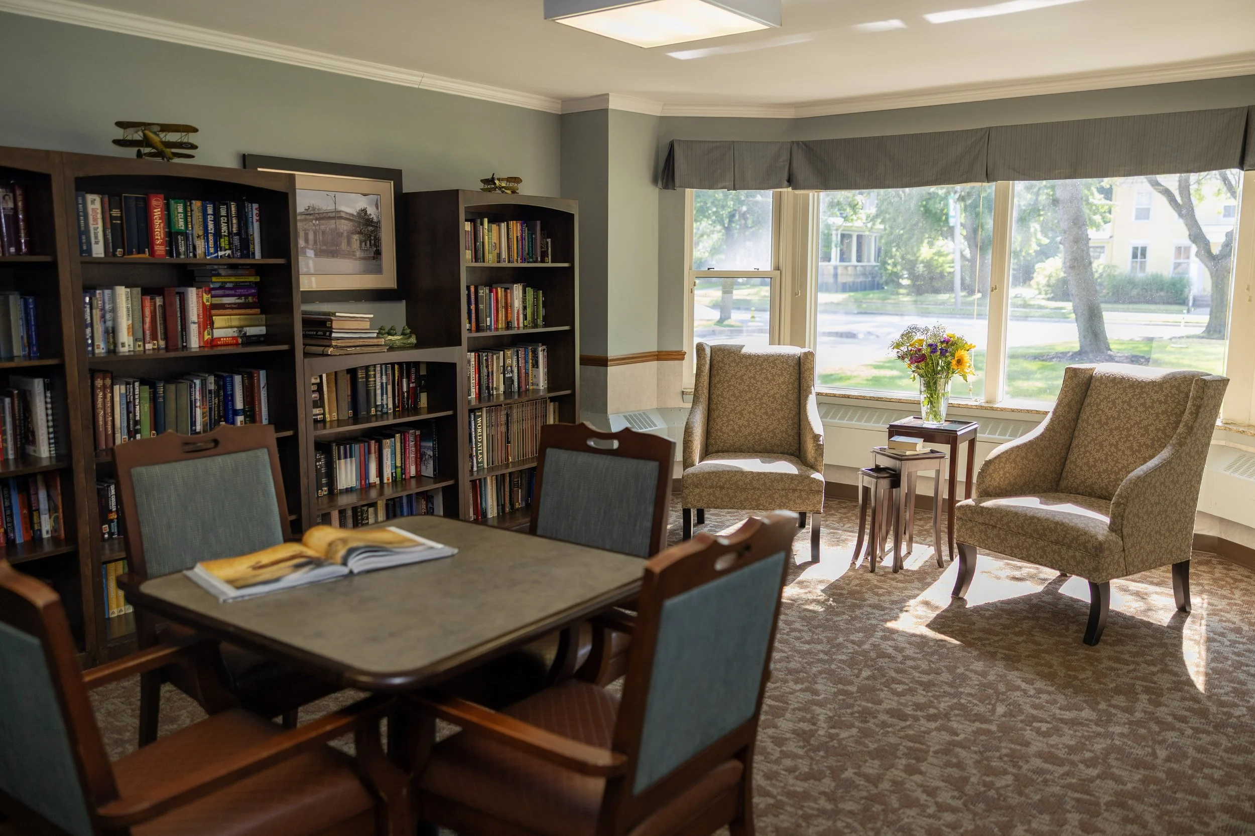 A cozy reading room with a wooden table, six upholstered chairs, and bookshelves filled with books. Large windows let in natural light, with a small table and chairs by the window, and a vase of flowers on the table beside two armchairs.