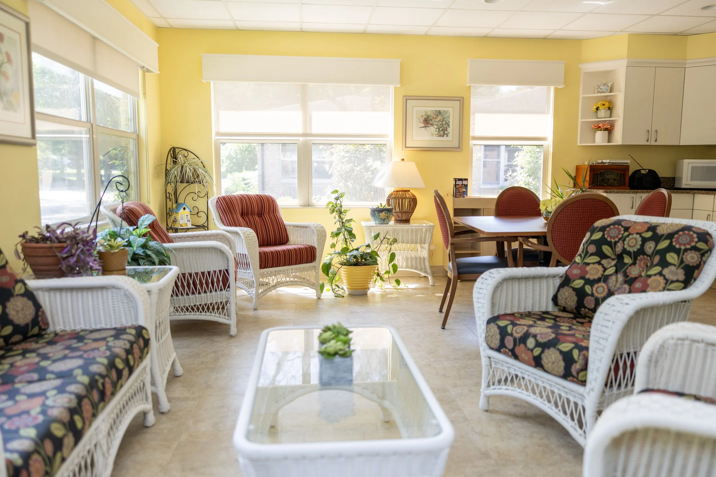 Bright, cozy room with yellow walls, wicker chairs with floral and striped cushions, a glass-top coffee table, potted plants, and a dining area with a wooden table and red chairs, sunlit through large windows.