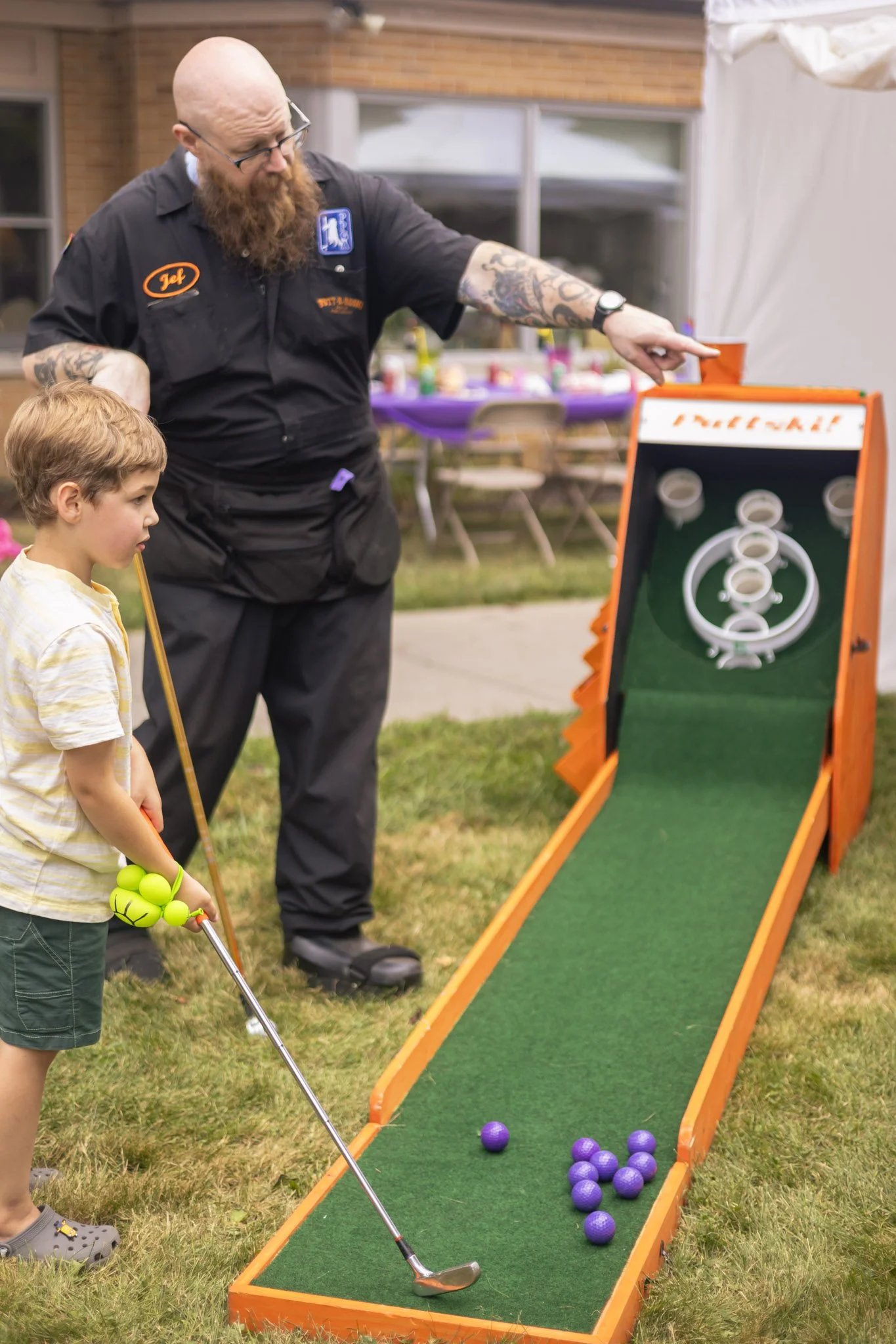 A bald man with a beard, wearing glasses and a tattooed arm, instructs a young boy holding a mini-golf club at a mini-golf game setup. Purple balls are on the green surface of the game. The setting is outdoors with a table and chairs in the backgroun