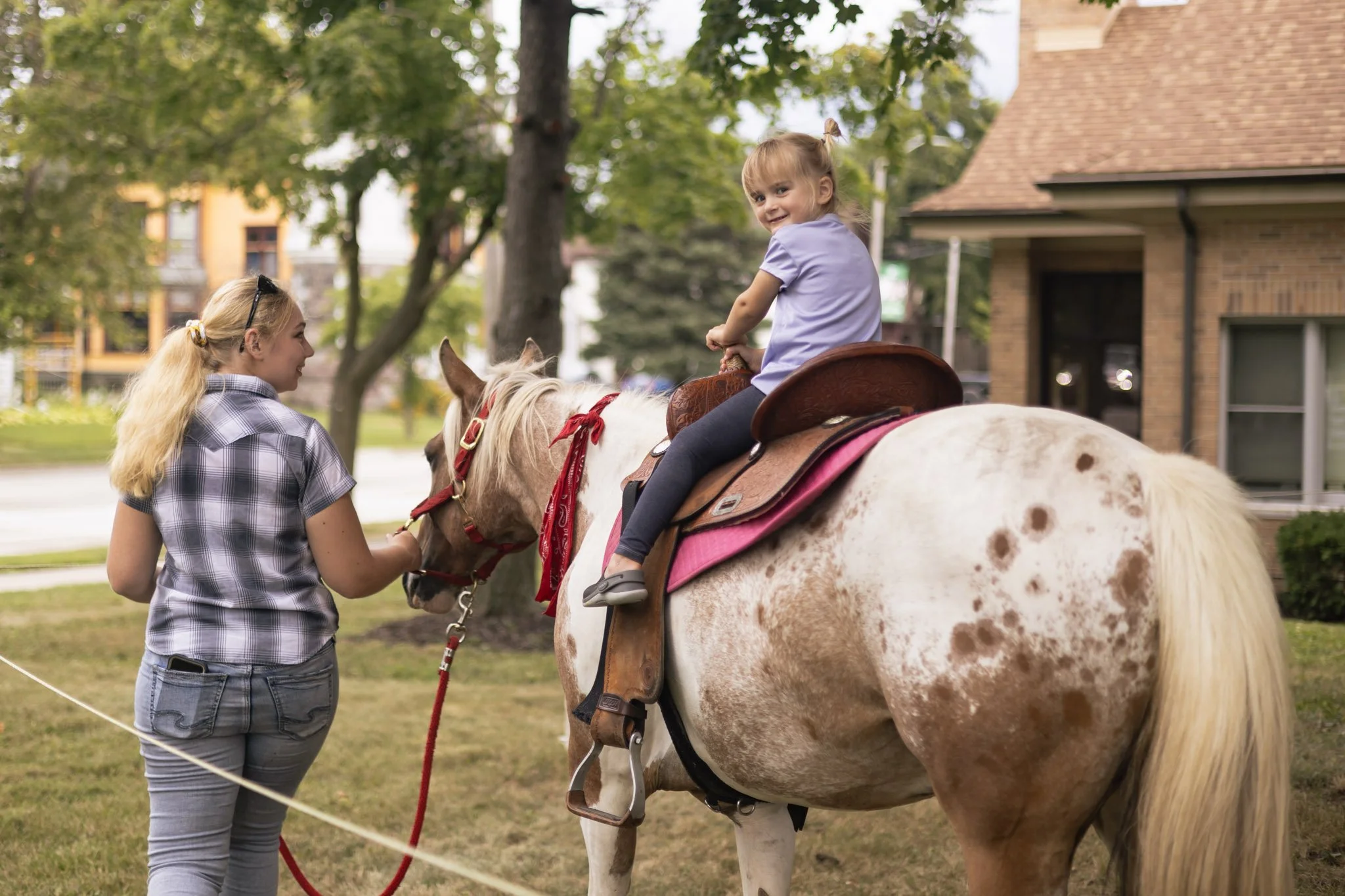 A young girl riding a spotted horse guided by a woman holding a red lead rope, outdoors near a house with trees.