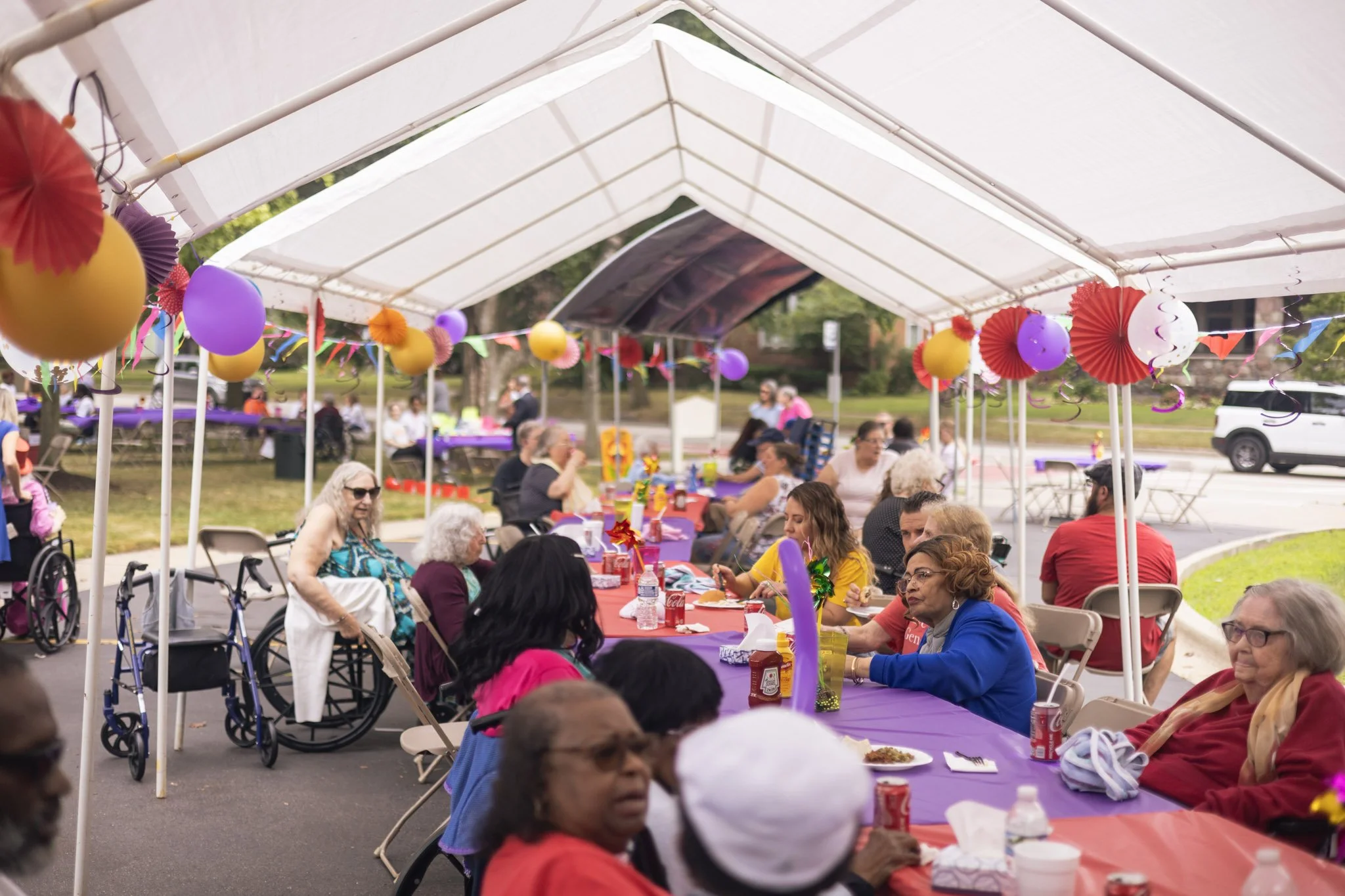 Outdoor gathering under a canopy with people seated at tables, decorated with colorful balloons, streamers, and tablecloths.
