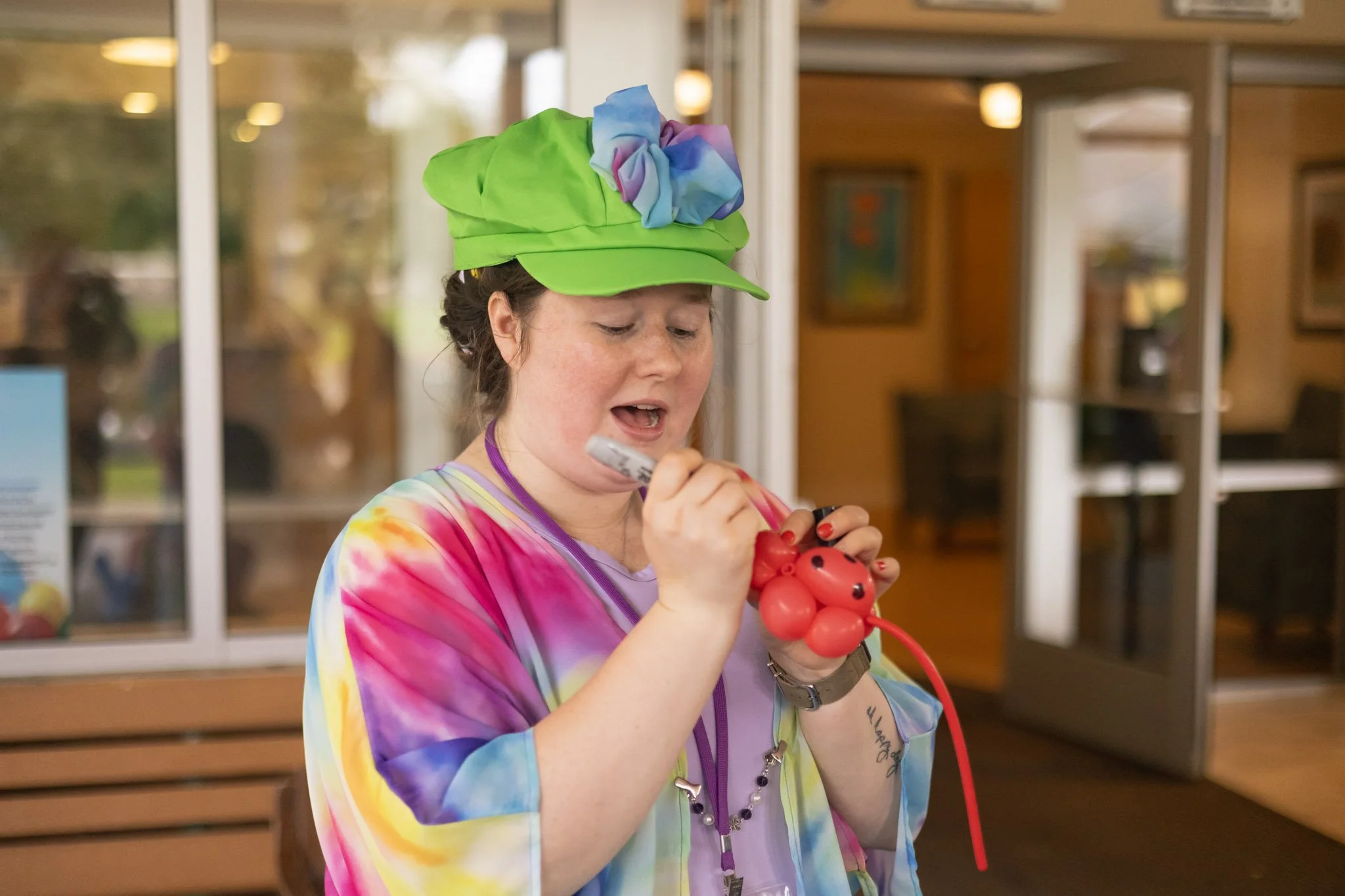 Person wearing a colorful outfit and green hat, inflating a balloon animal, while holding a marker.