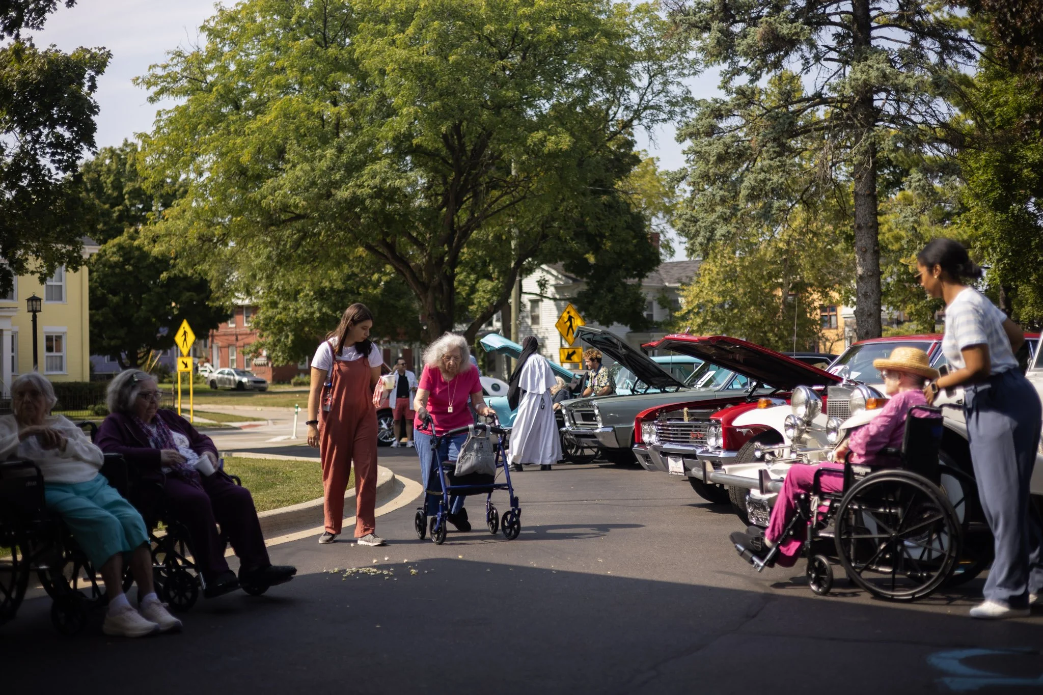 Elderly women sitting in wheelchairs and walking with assistance at an outdoor car show on a sunny day, with vintage cars on display and trees in the background.