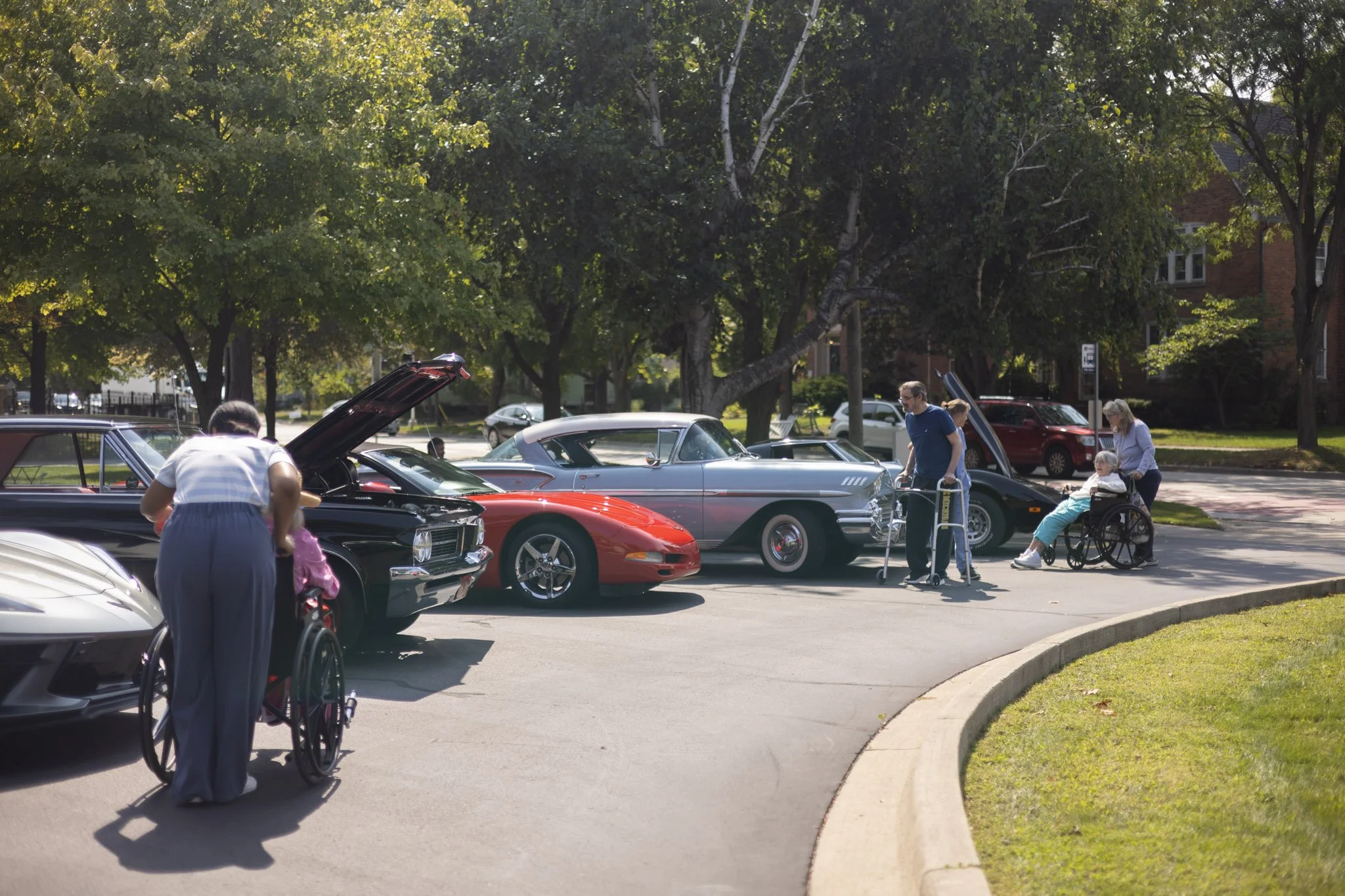 People visiting a classic car show in a park, standing and sitting near vintage cars on display, with trees and houses in the background.