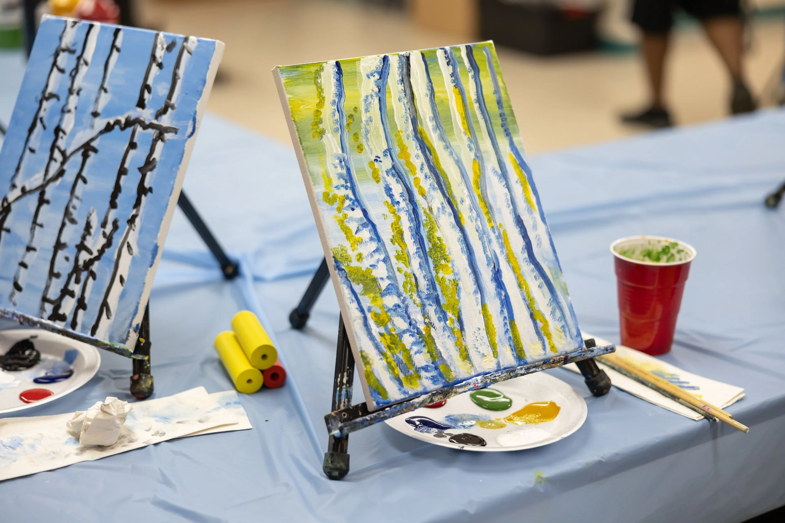 Two small easels hold canvases with abstract paintings of trees with white and black trunks and green, white, yellow, and blue foliage, set on a table covered with a blue plastic tablecloth, surrounded by paint supplies and a red cup.