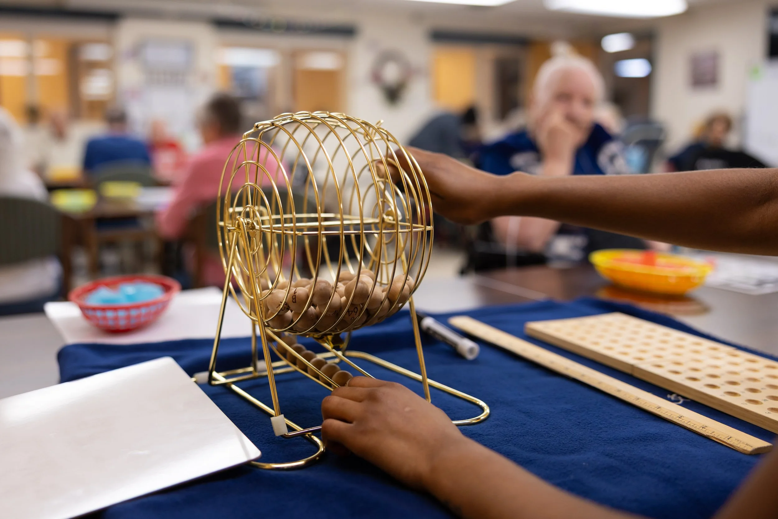 A person's hand is pulling a bingo ball from a gold bingo cage, with bingo cards and a marker on a table in the activity room.