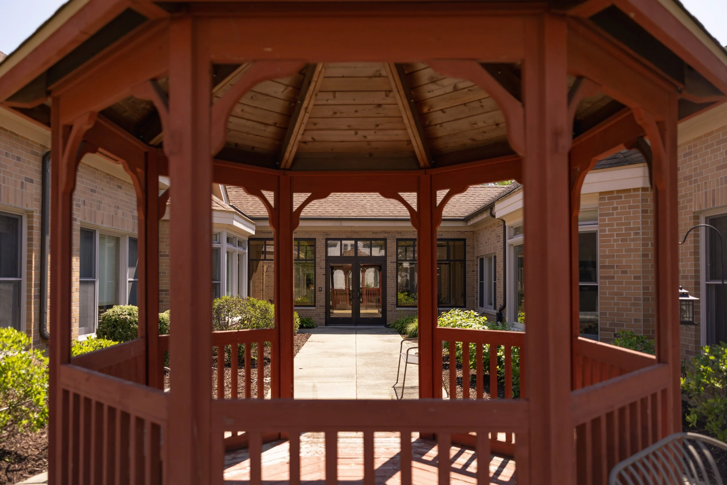 View of a red wooden gazebo in the backyard of a house with brick exterior, large windows, and garden plants.
