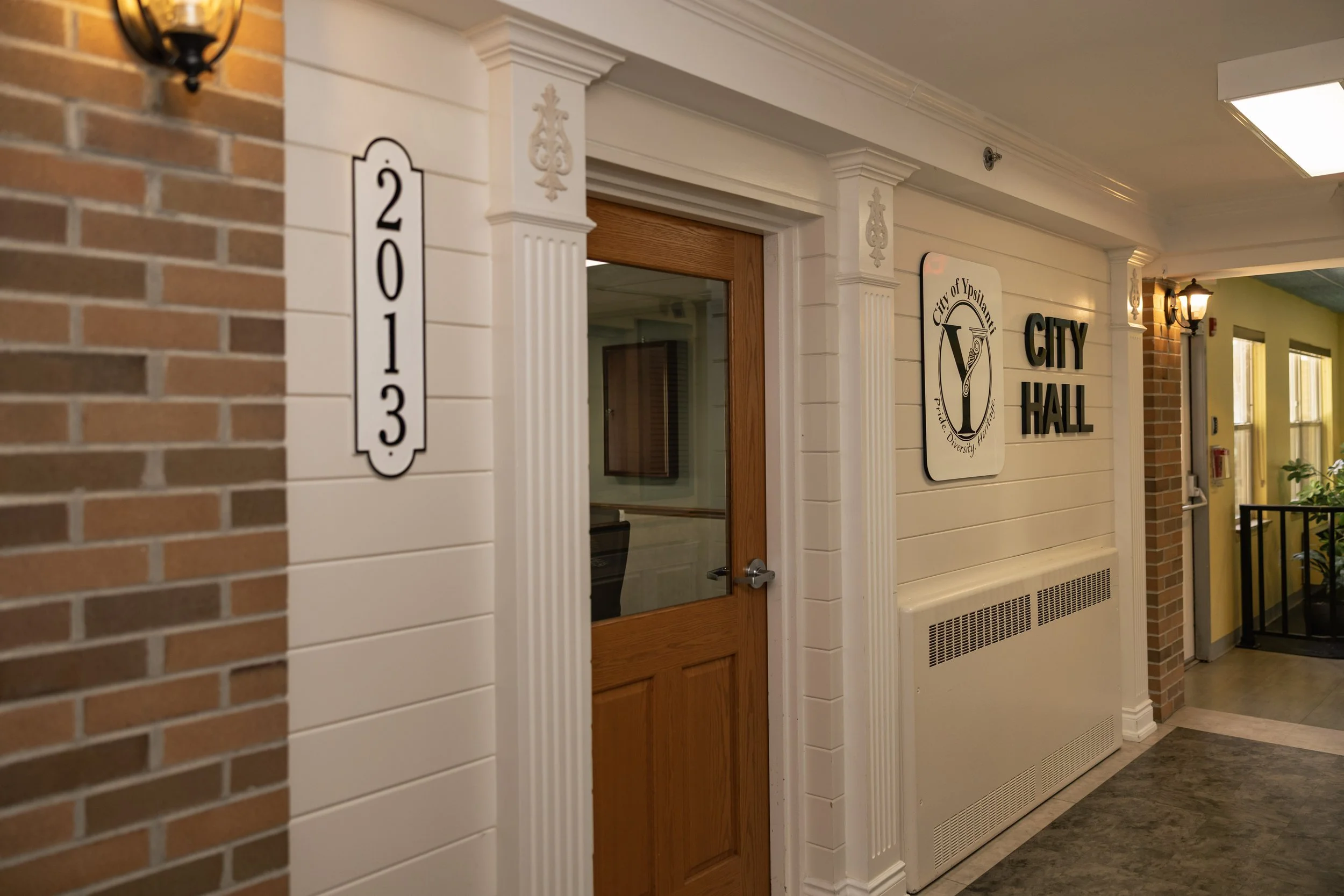 Interior of a city hall with a door, signs indicating the city of Ypsilanti, and room number plaque 2013. A reference to when the building addition was added to the facility.