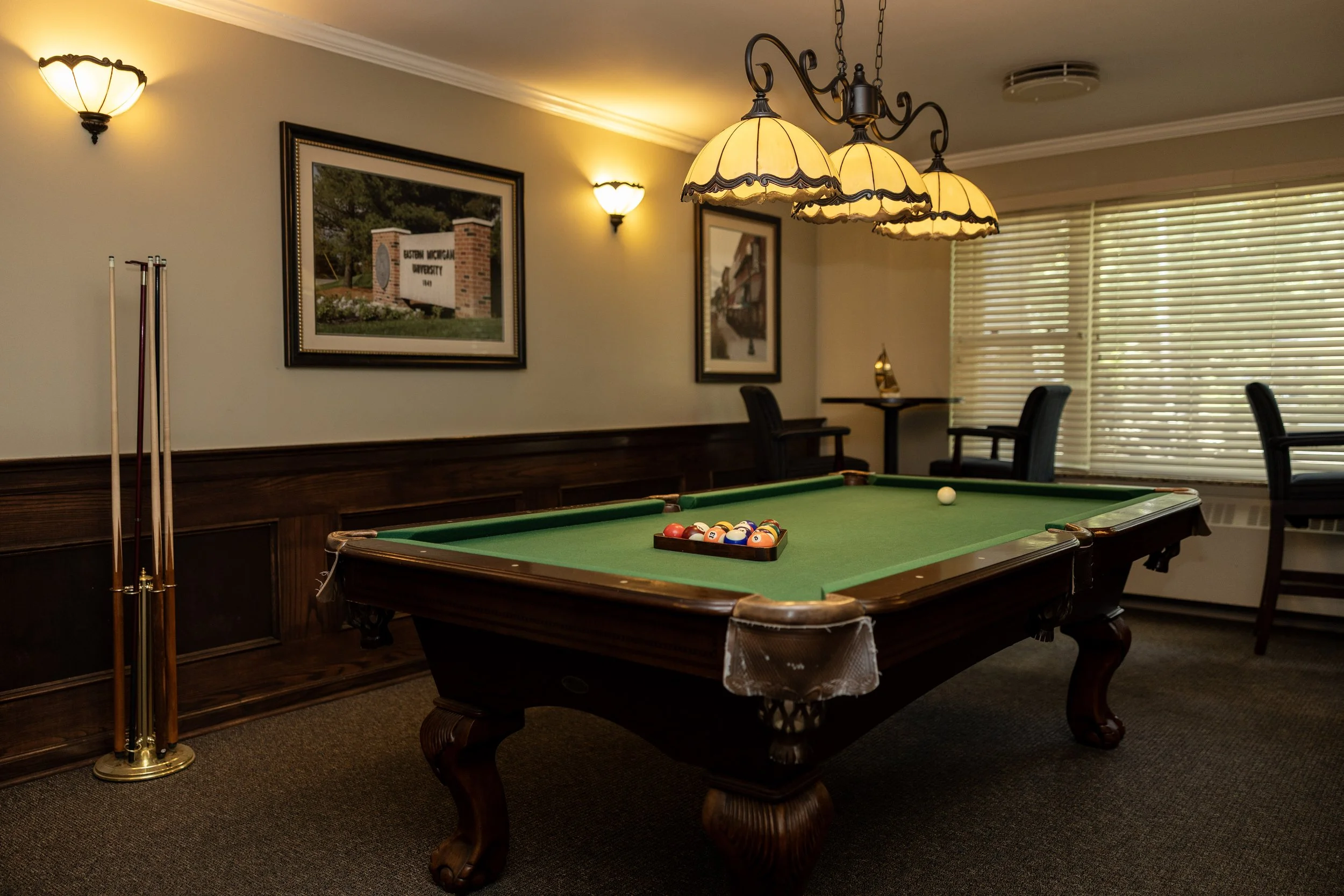 An indoor billiards room with a pool table, pool balls racked, pool cue sticks, wall-mounted lights, framed pictures, window blinds, and chairs.