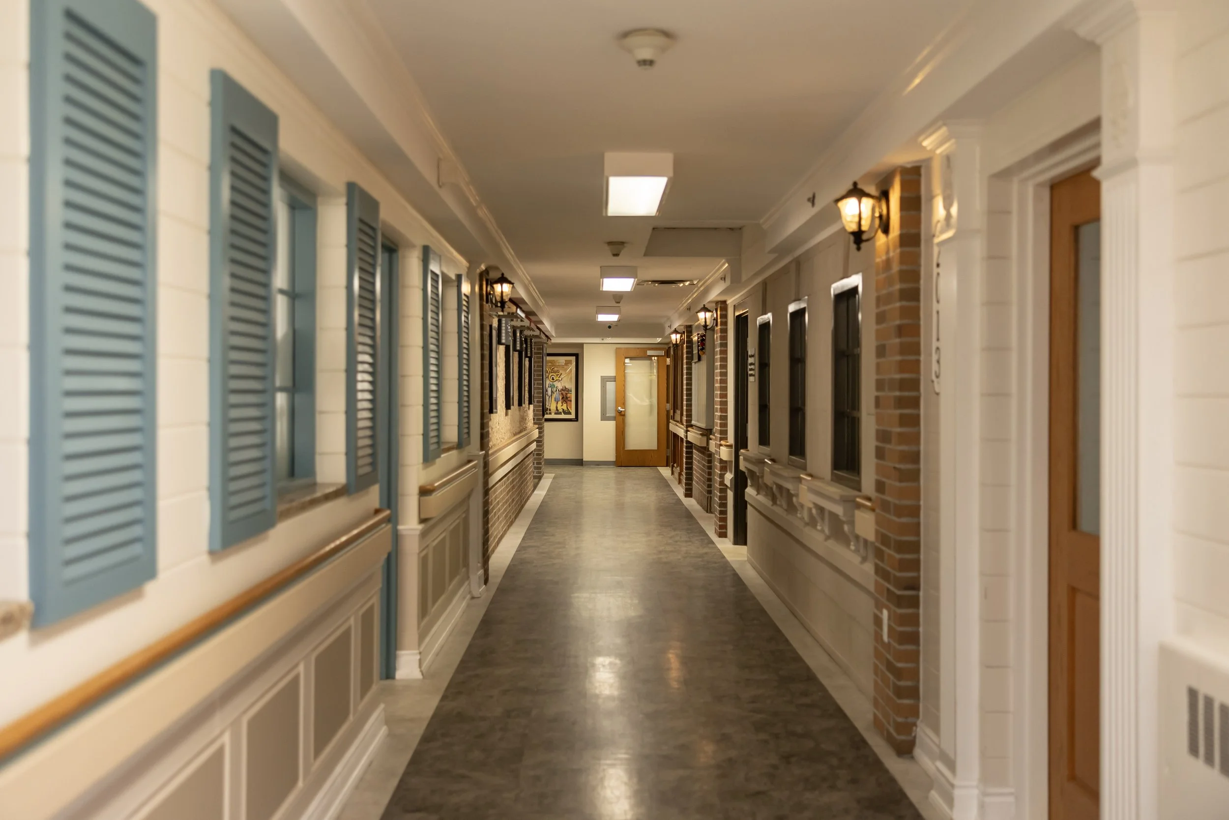 Empty indoor corridor with beige walls, blue window shutters, brick accents, and wall-mounted lantern lights.