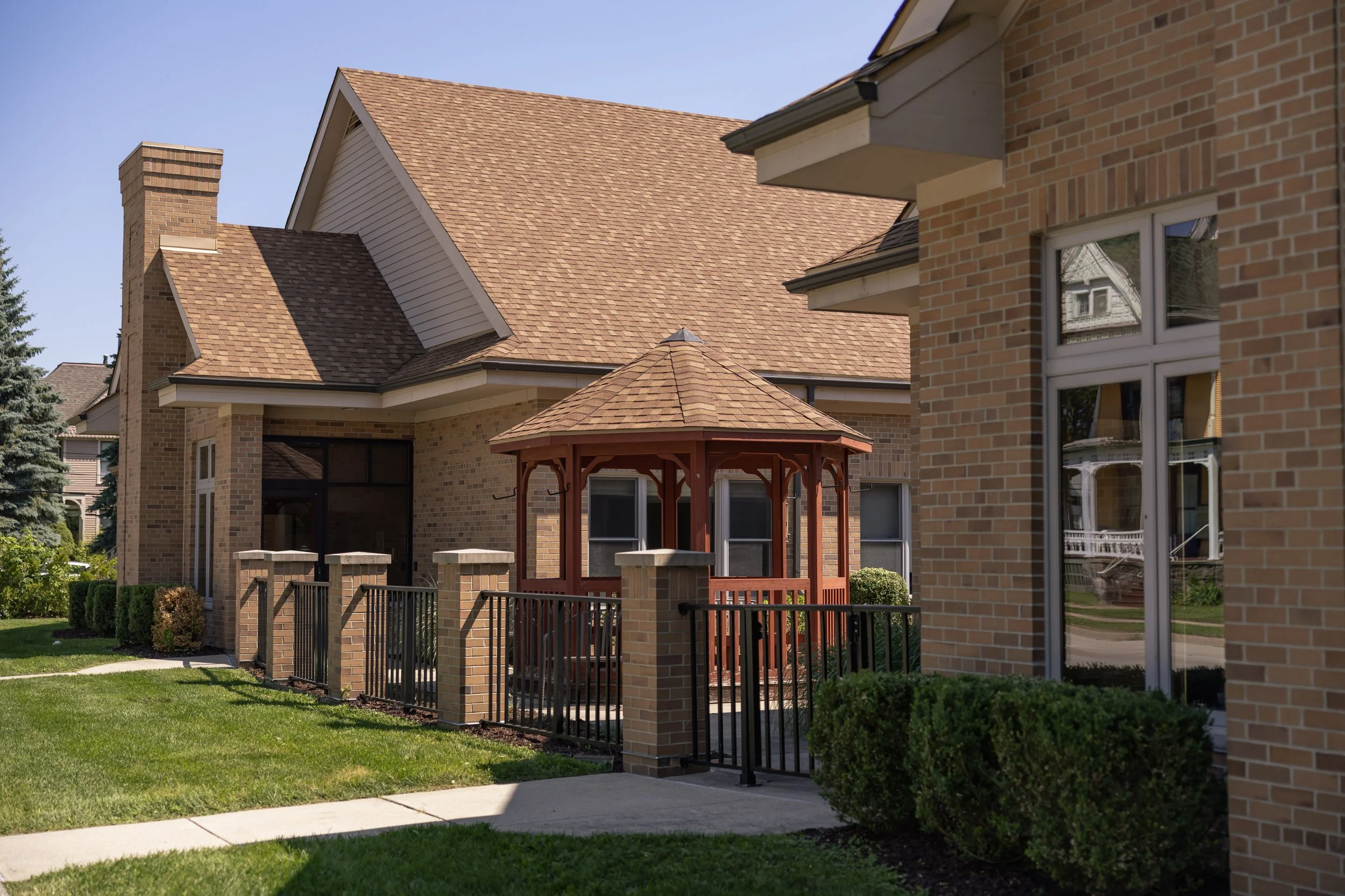A brick residential house with a brown shingled roof, a small wooden gazebo, a fenced patio area, and a green lawn.
