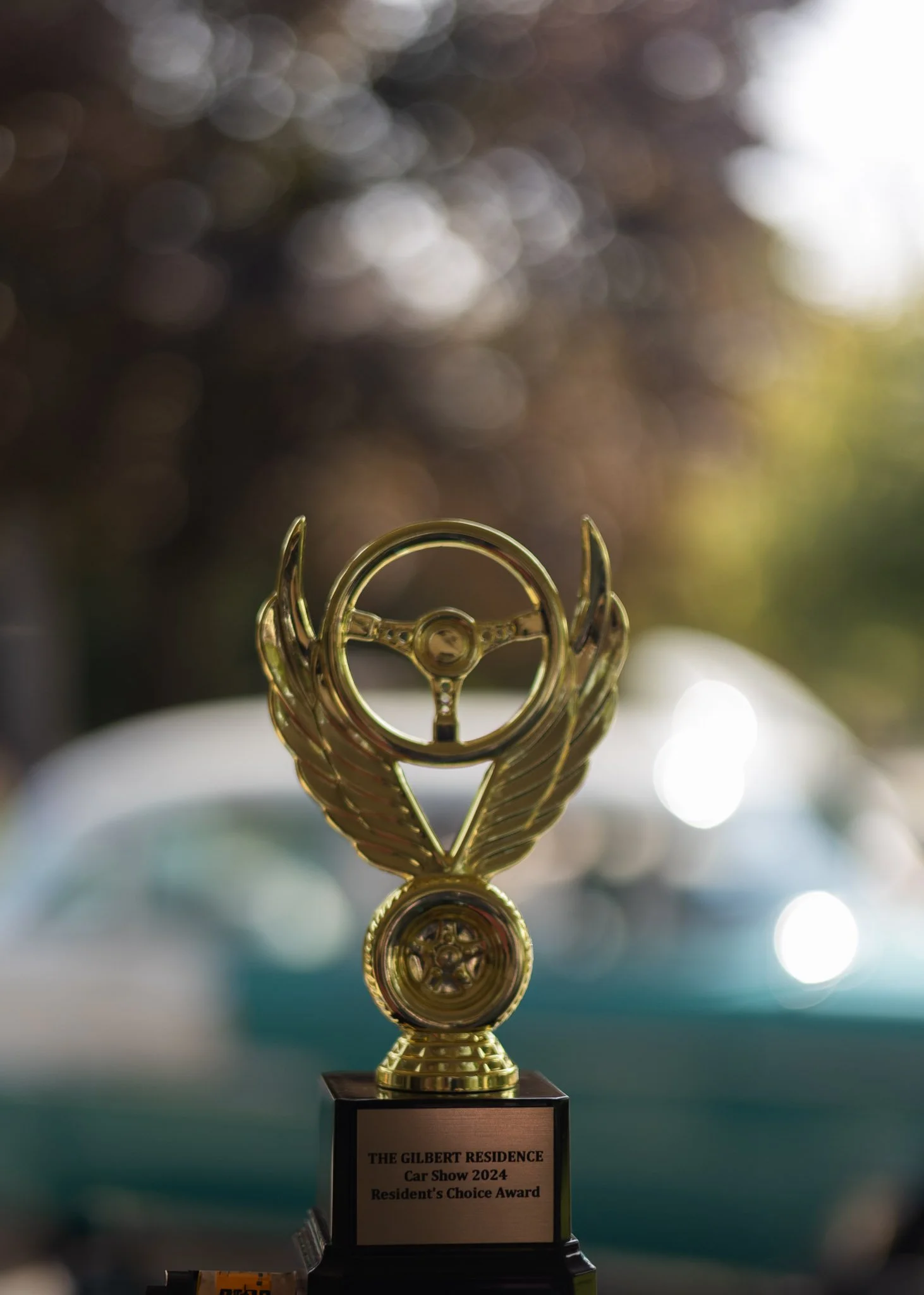A gold trophy with a steering wheel and winged design, awarded at a car show, with a background of blurred sunlight and trees.
