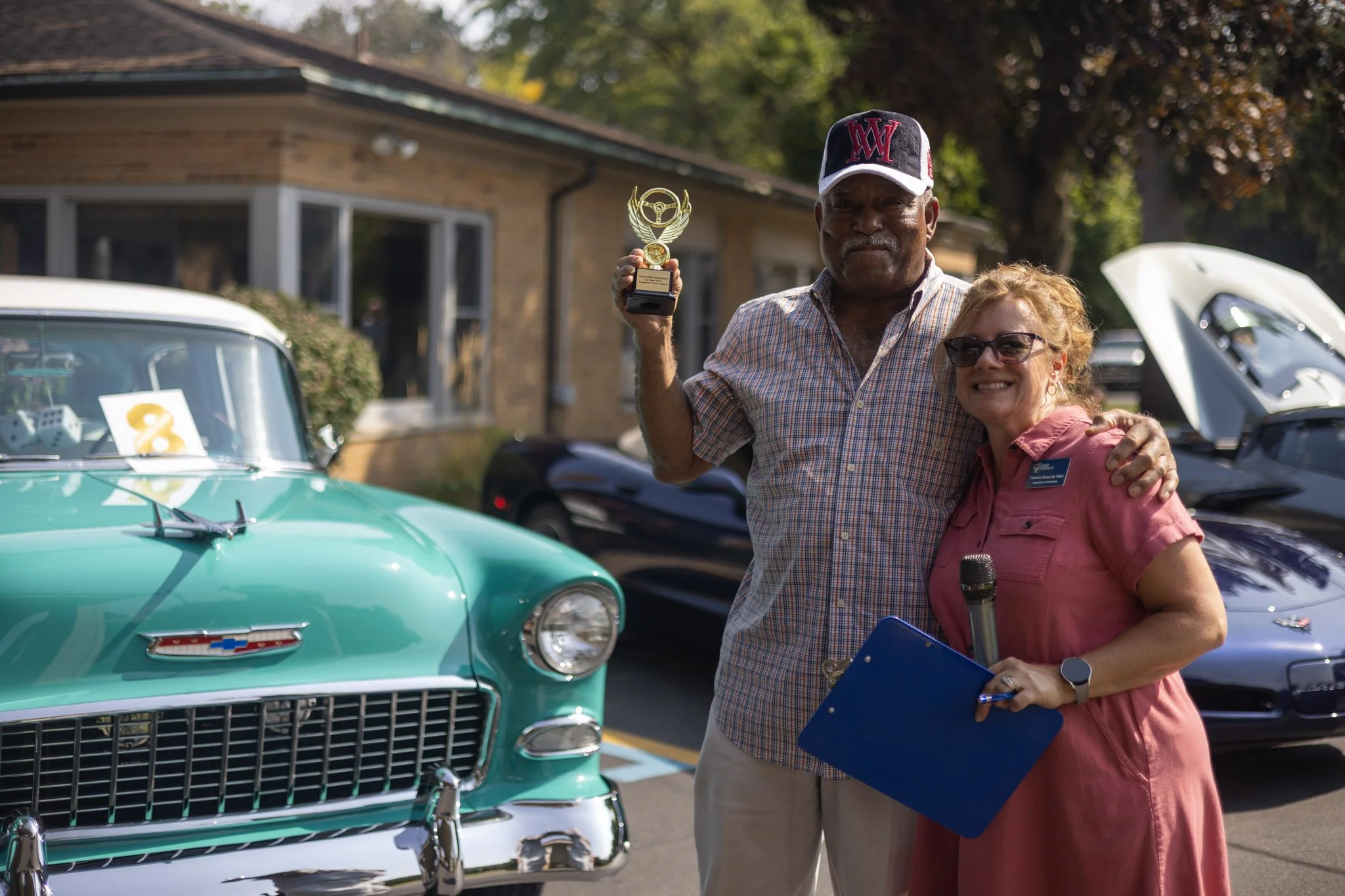 A man holding a trophy and a woman holding a microphone and clipboard, standing next to a vintage teal Chevrolet, at an outdoor car show with a black sports car in the background.