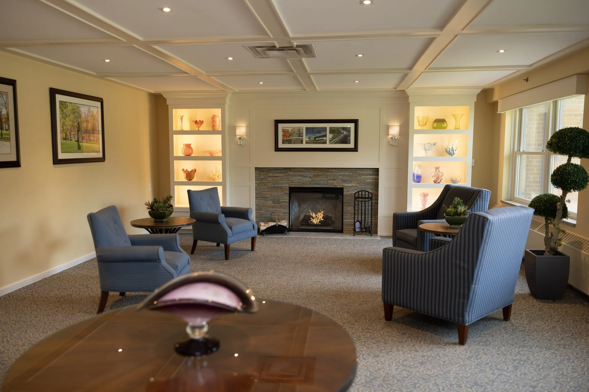 Living room with a fireplace, upholstered armchairs, glass shelves with decorative glassware, and framed artwork.