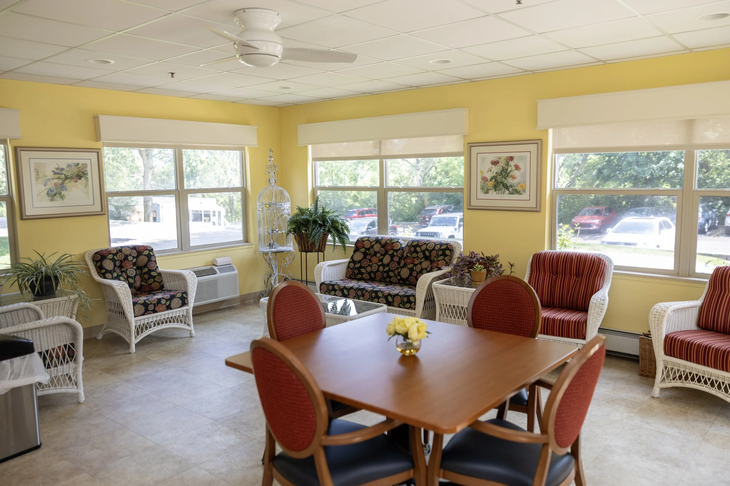 Bright yellow room with large windows, colorful wicker armchairs, a wooden dining table with red chairs, framed floral artwork, and indoor plants.