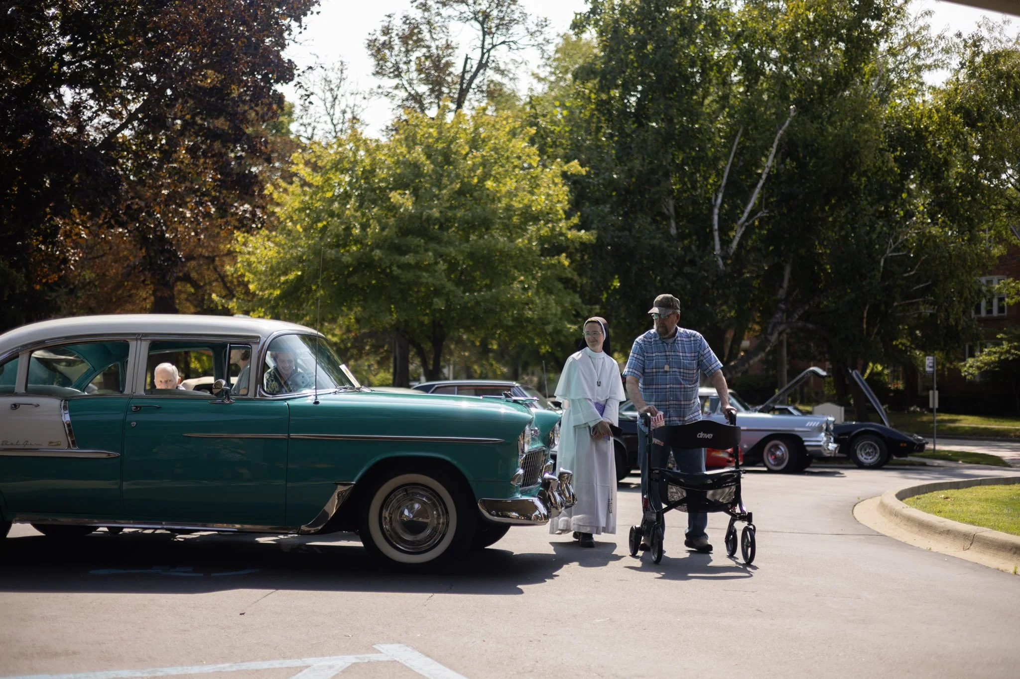 A man with a walker and a woman dressed as a nun talking to an elderly person in a vintage car at a car show in a park with trees.