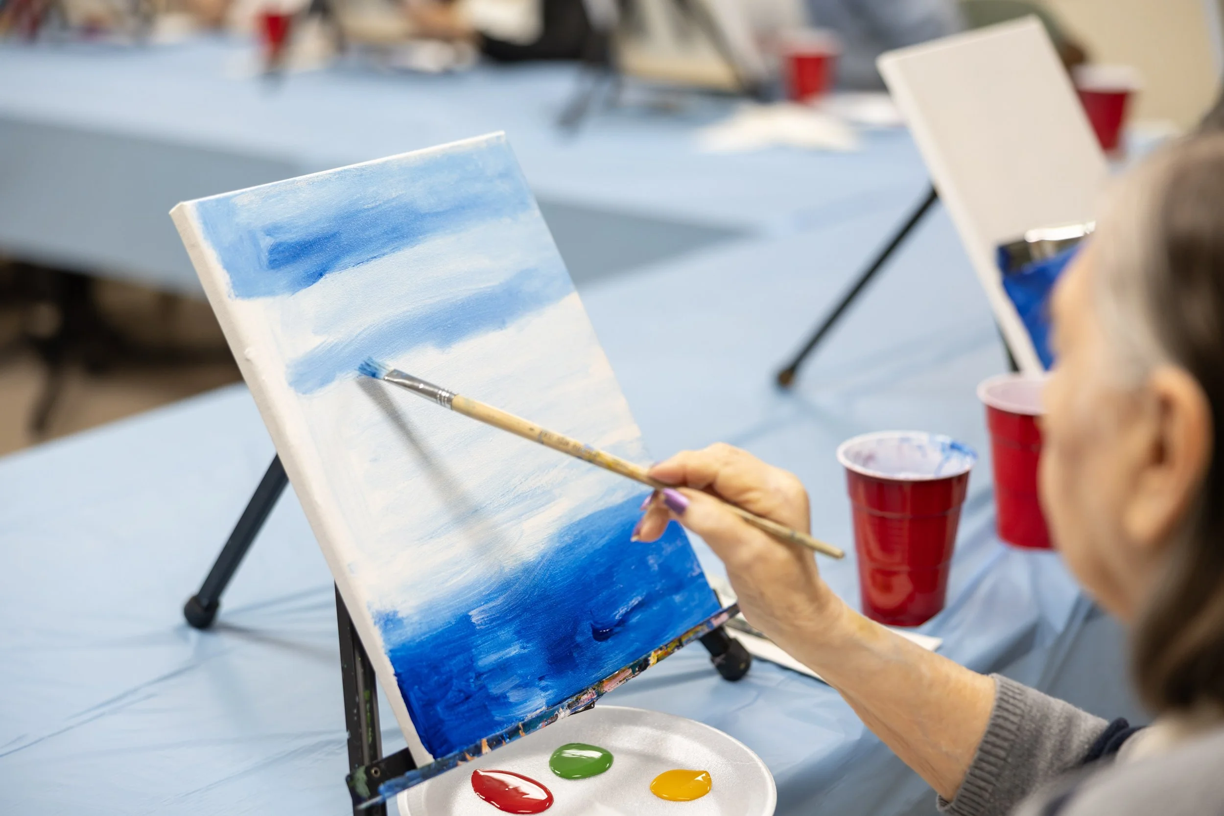 An elderly woman painting a seascape with blue sky and ocean on a canvas at an art class, with red and blue cups and paint on the table.