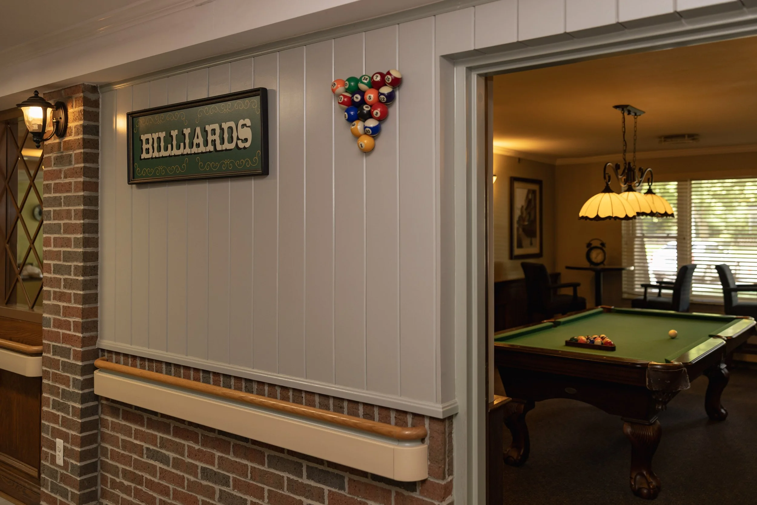 Interior of a recreational room with a pool table, hanging lamps, and wall decor, viewed through an open doorway from a hallway with a 'Billiards' sign and a wall-mounted set of billiard balls arranged in a triangle.