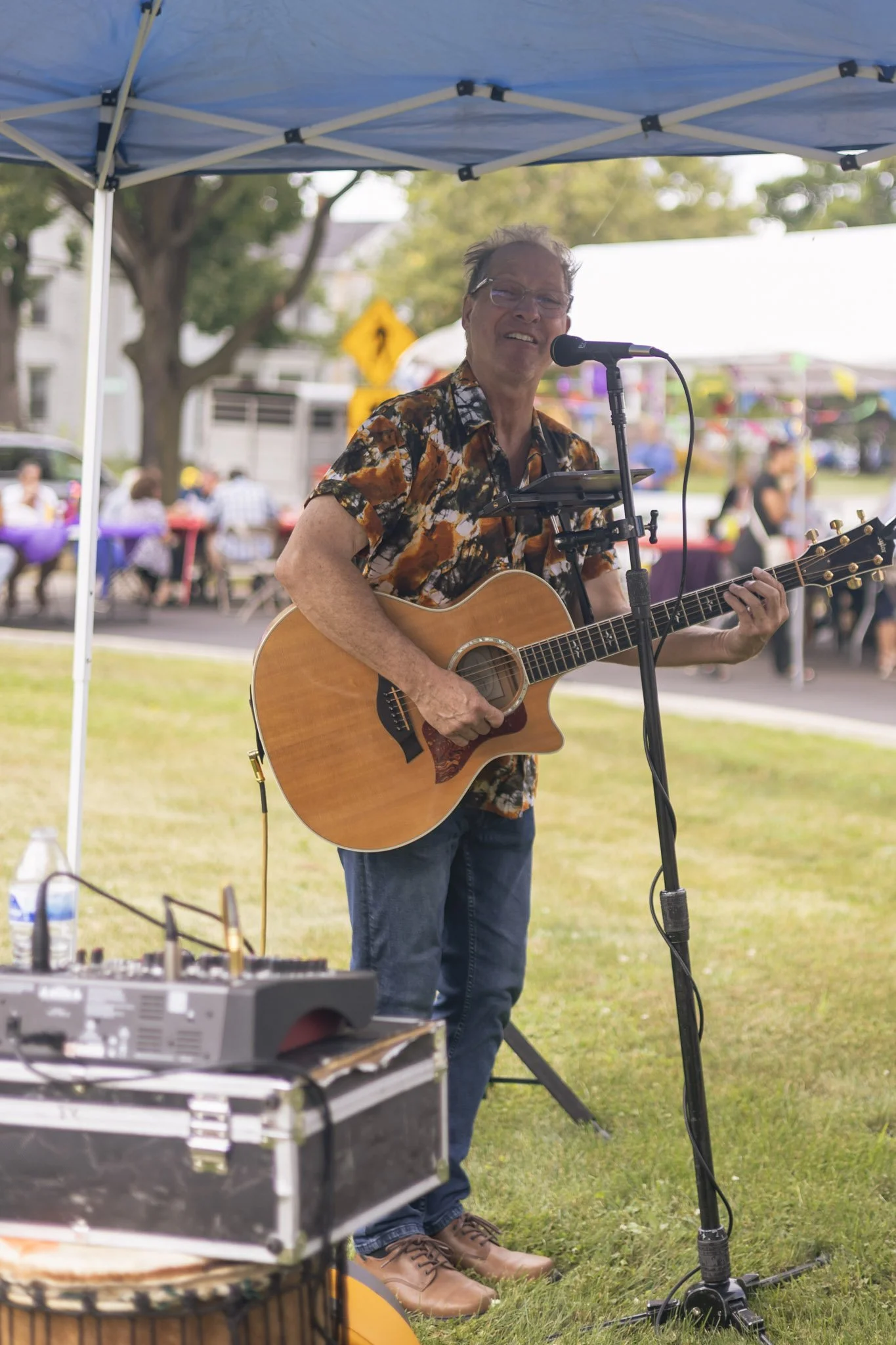 Musician performing outdoors with a guitar and microphone under a blue canopy.