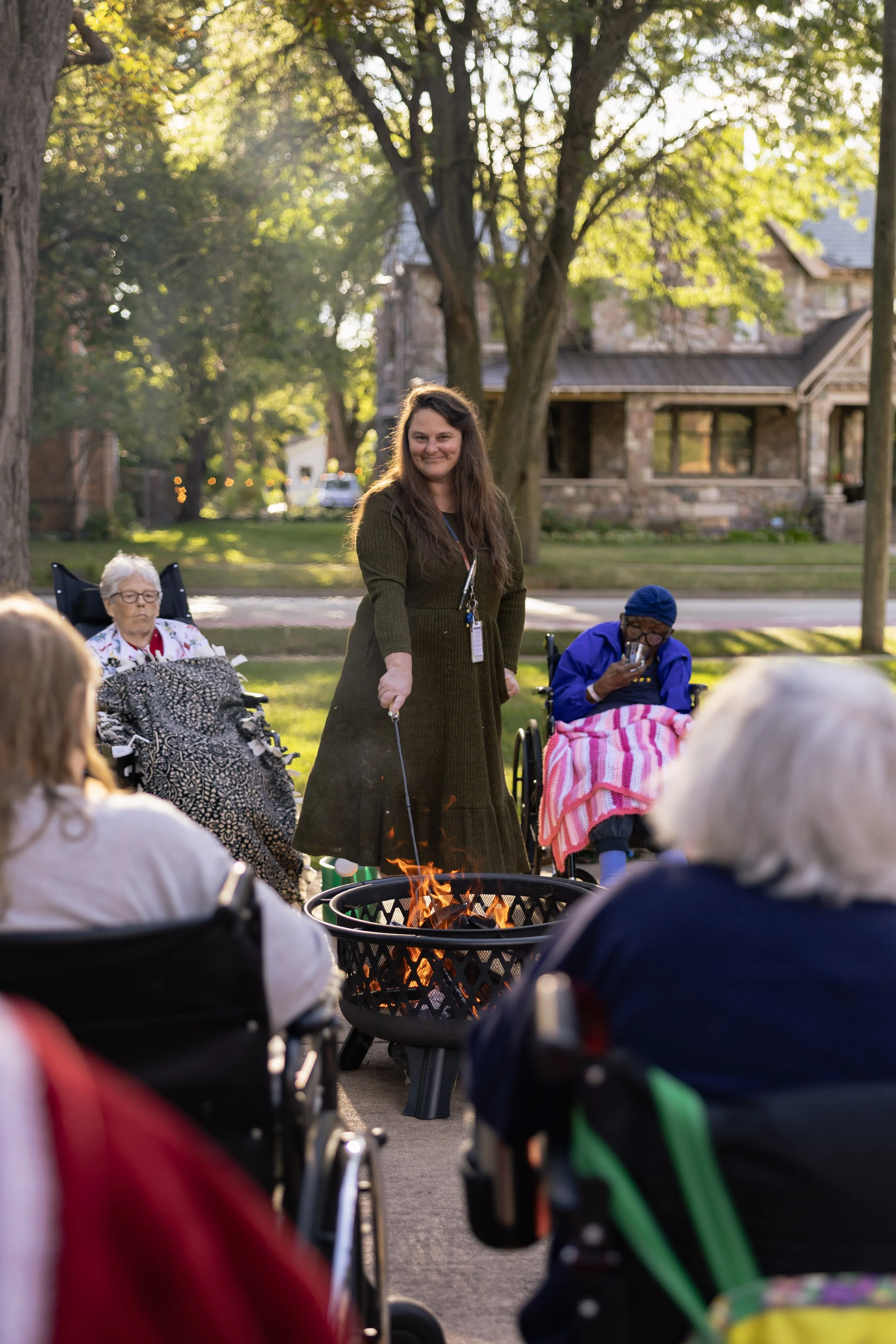 Maureen standing and roasting marshmallows over a firepit outdoors, surrounded by elderly women in wheelchairs, during what appears to be a social gathering on a sunny afternoon.