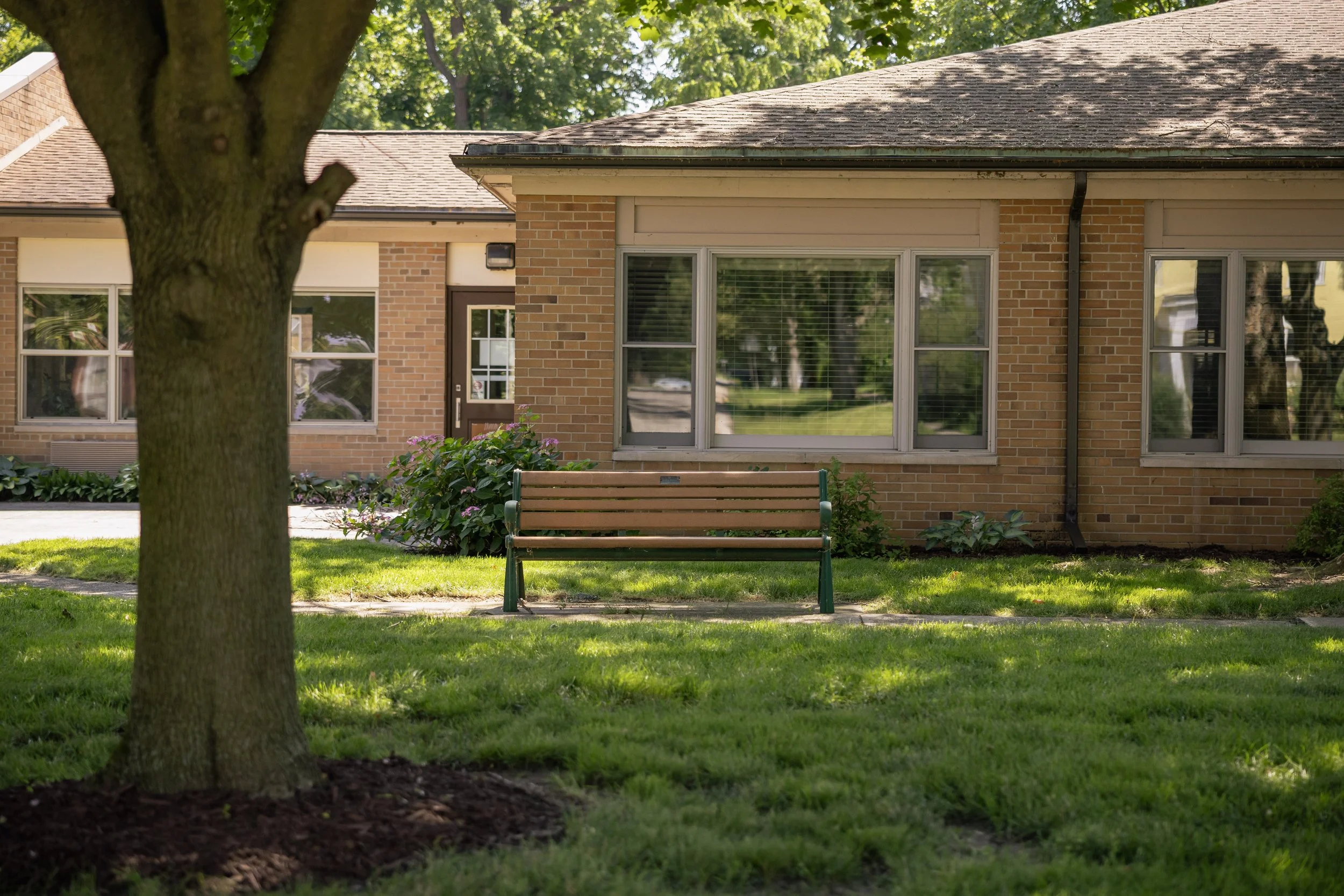 A brick building with large windows, a brown door, and a brown roof, surrounded by a grassy lawn, trees, and a wooden bench in front of the building.
