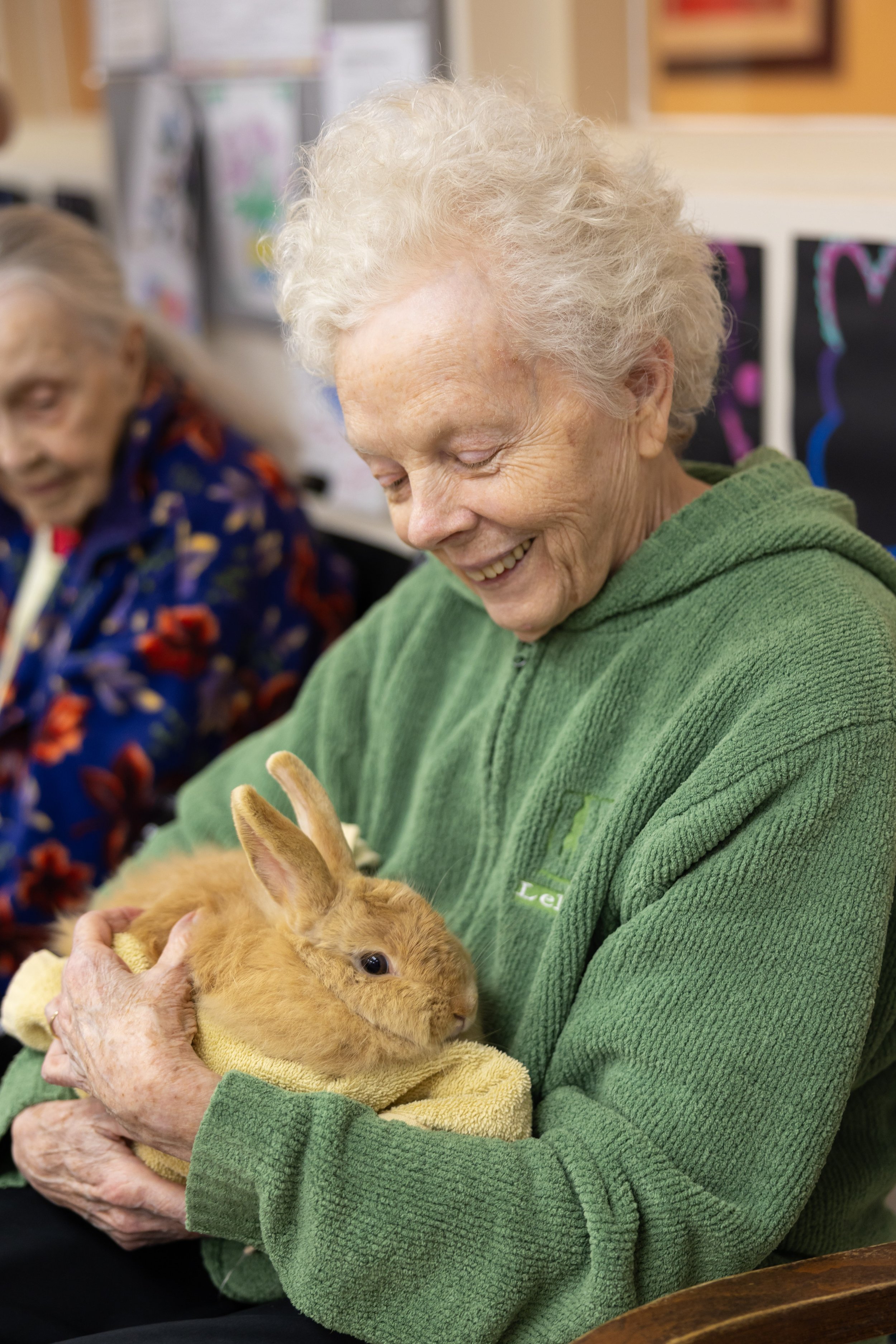 An elderly woman in a green fleece jacket holding a fluffy, light brown rabbit wrapped in a yellow towel, smiling happily at the rabbit.