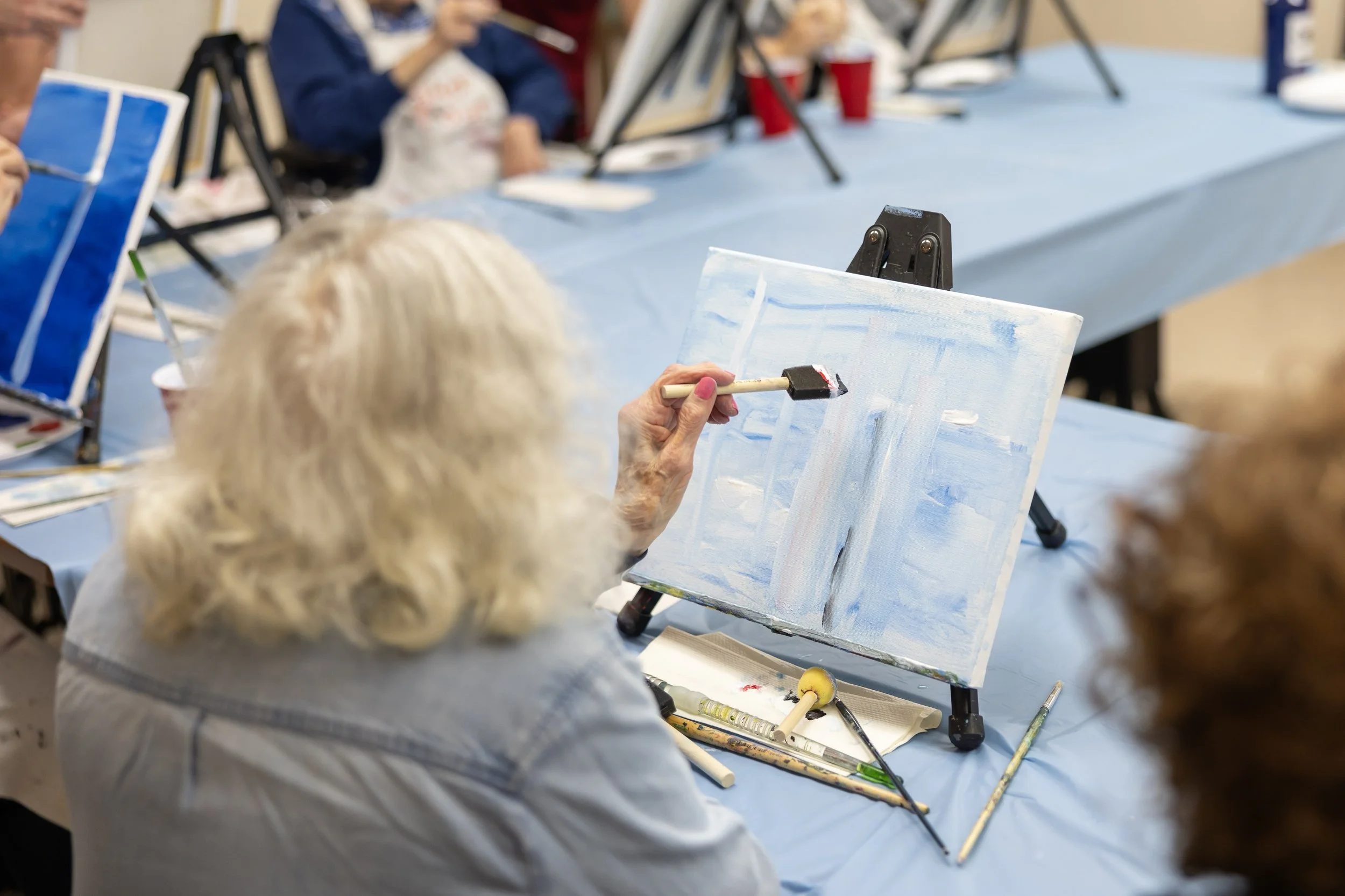 Older woman painting an abstract landscape on a small canvas during a group art class, with other participants visible in the background.