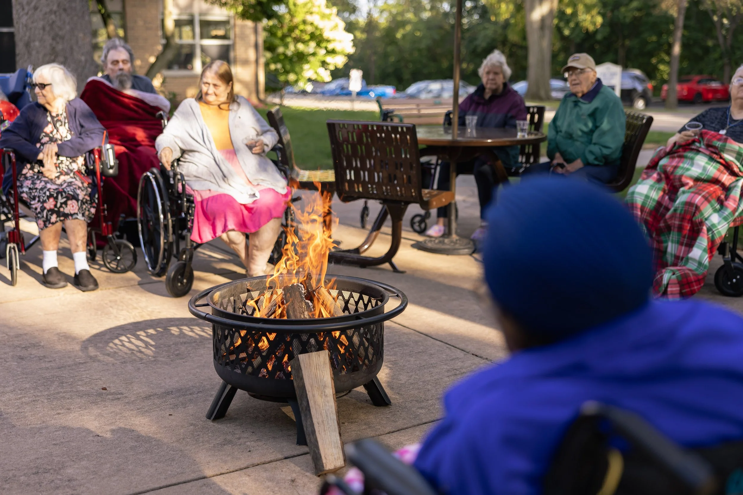 Group of elderly people sitting outdoors around a campfire on a patio, some in wheelchairs and some in chairs, with trees and parked cars in the background.