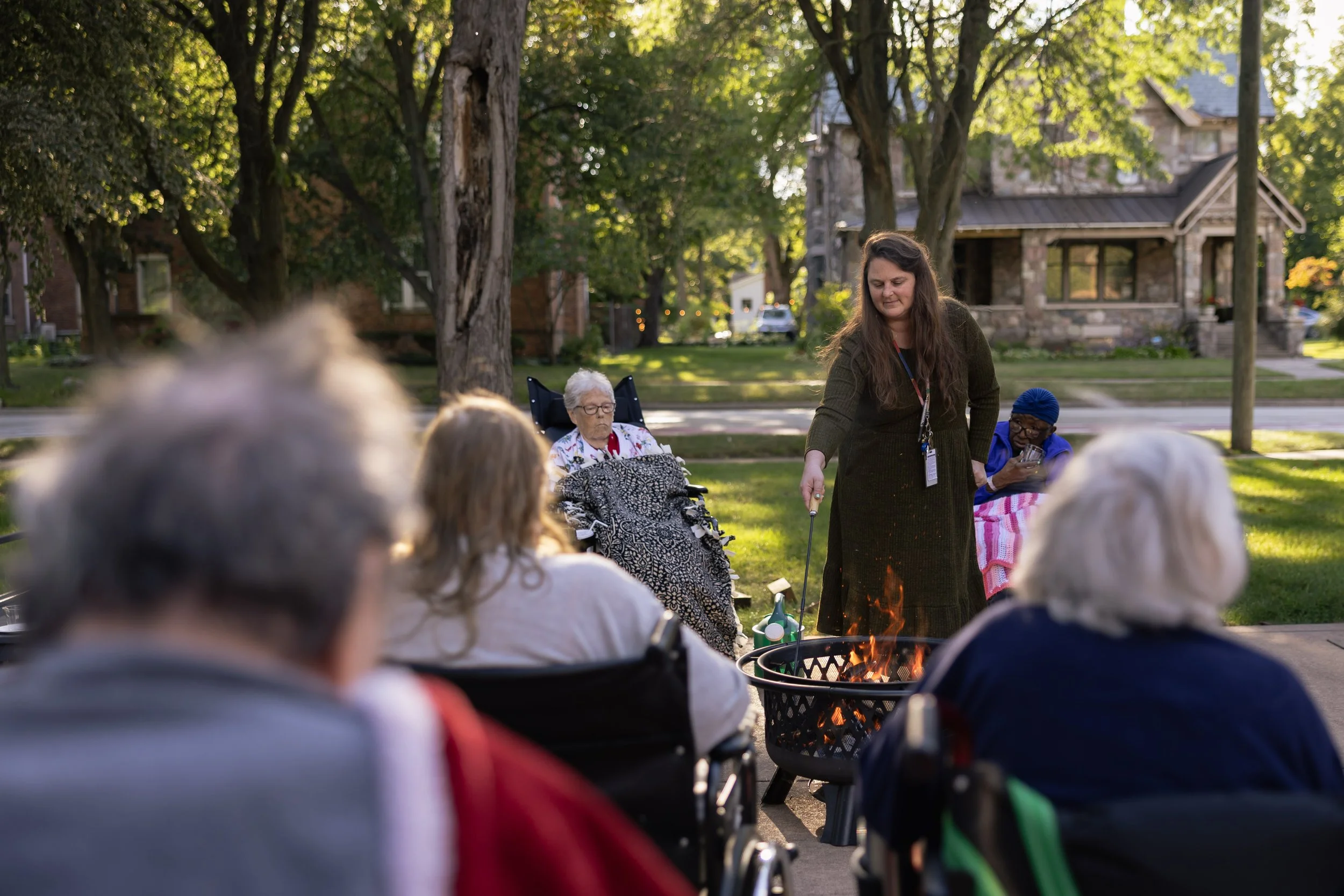 A group of elderly people gathered outdoors in a park for a social activity, with some in wheelchairs, while Maureen tends to a small fire in a fire pit on a sunny day.