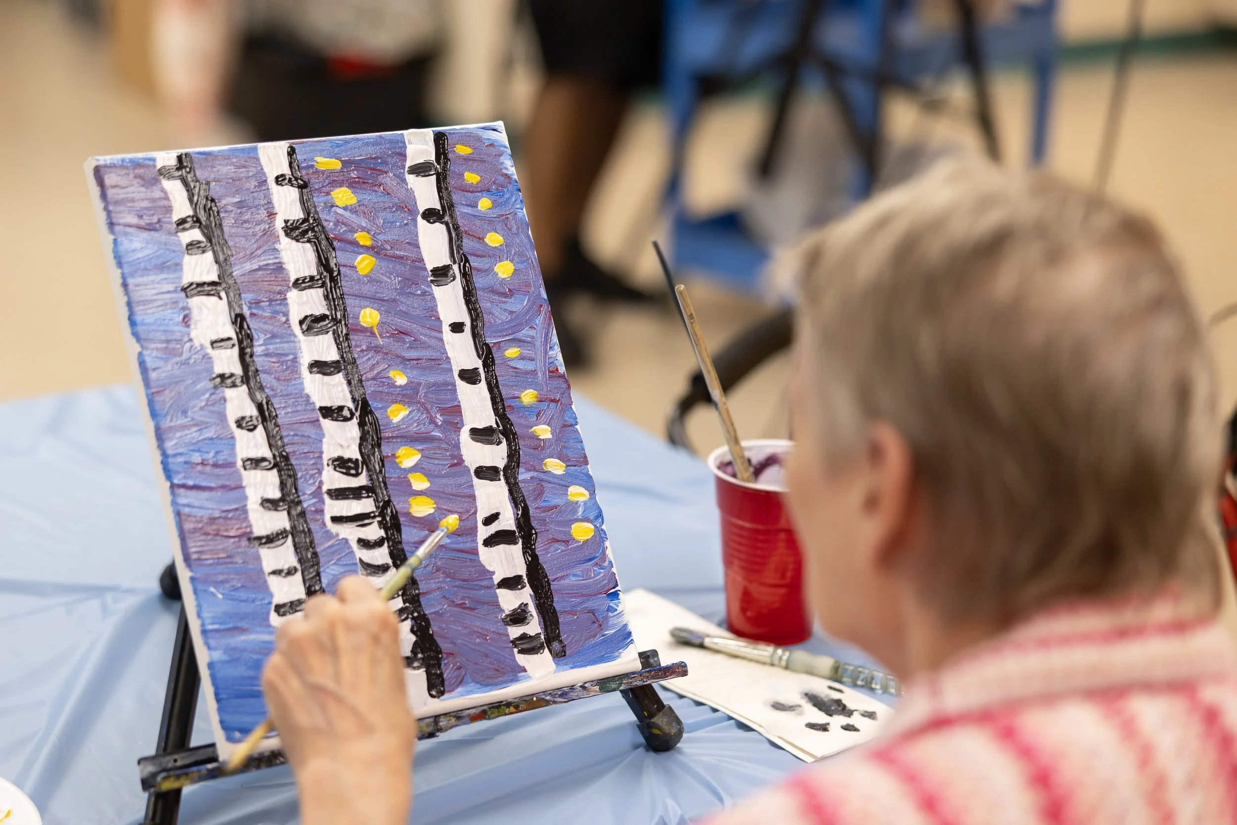 An elderly person painting an abstract landscape with vertical black and white lines and yellow dots on a blue-purple background at an art class.