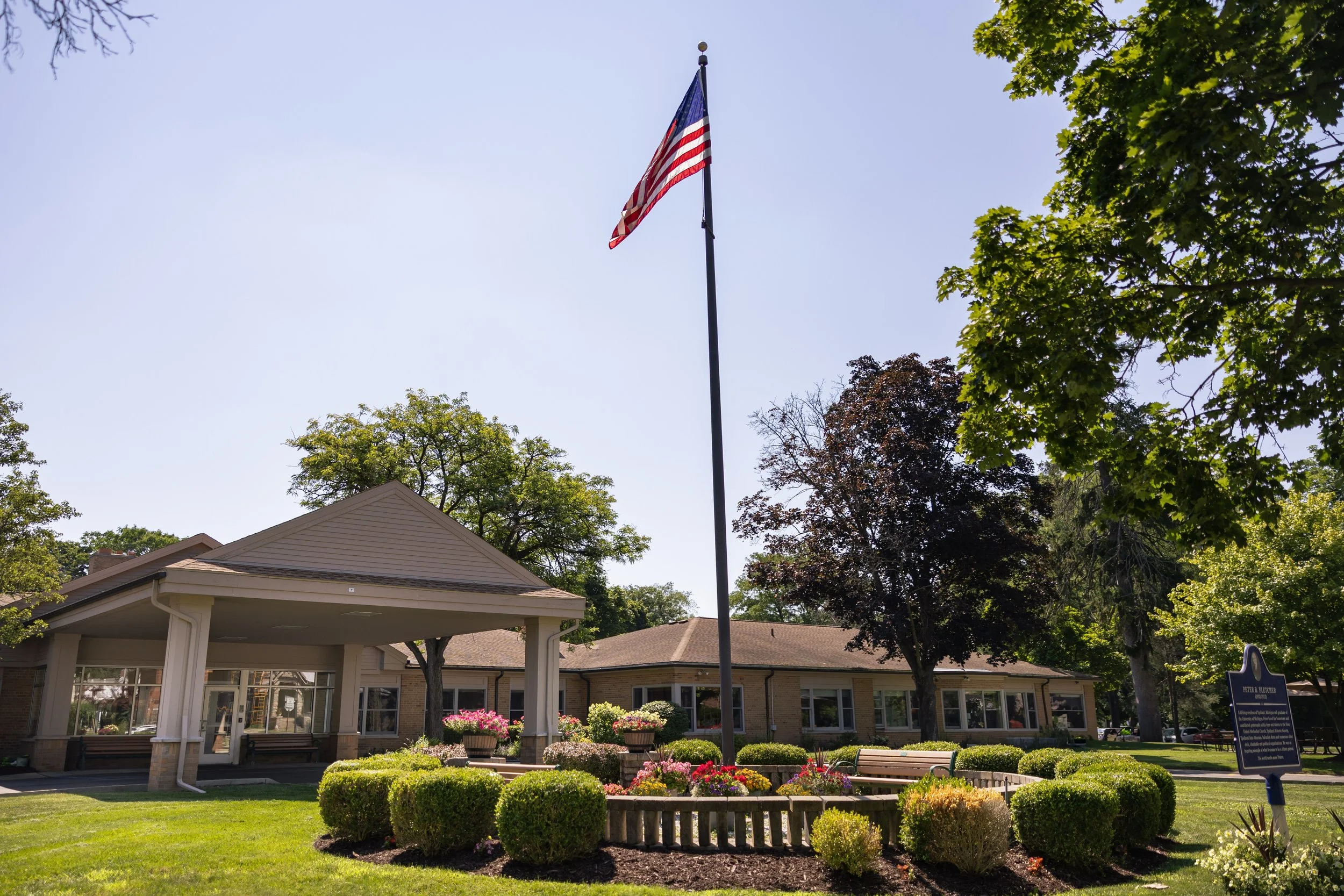 A flagpole with an American flag in front of a building surrounded by trees and landscaped bushes and flowers.