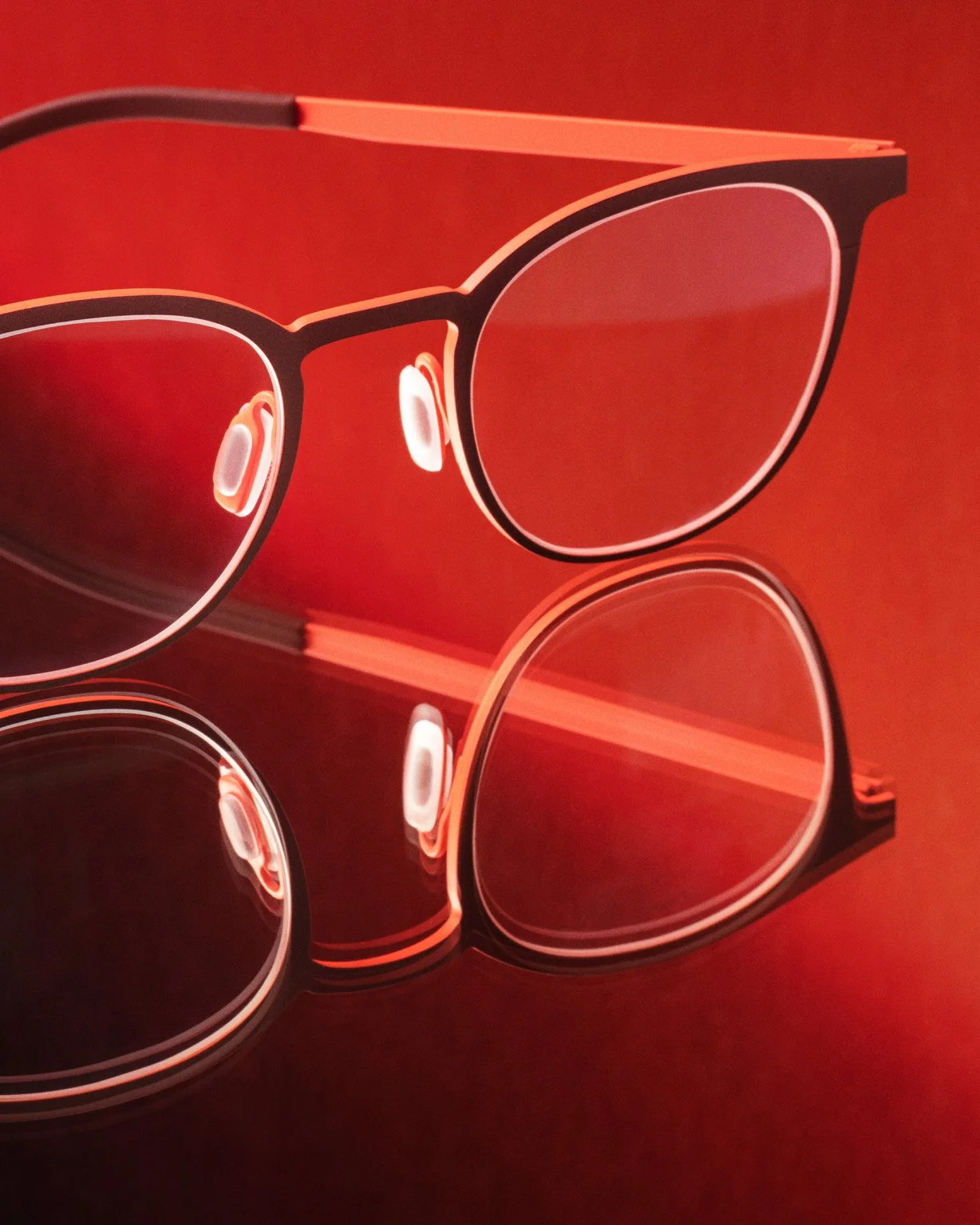Stylish eyeglasses with red frames and clear lenses reflecting on a shiny surface, set against a red background.