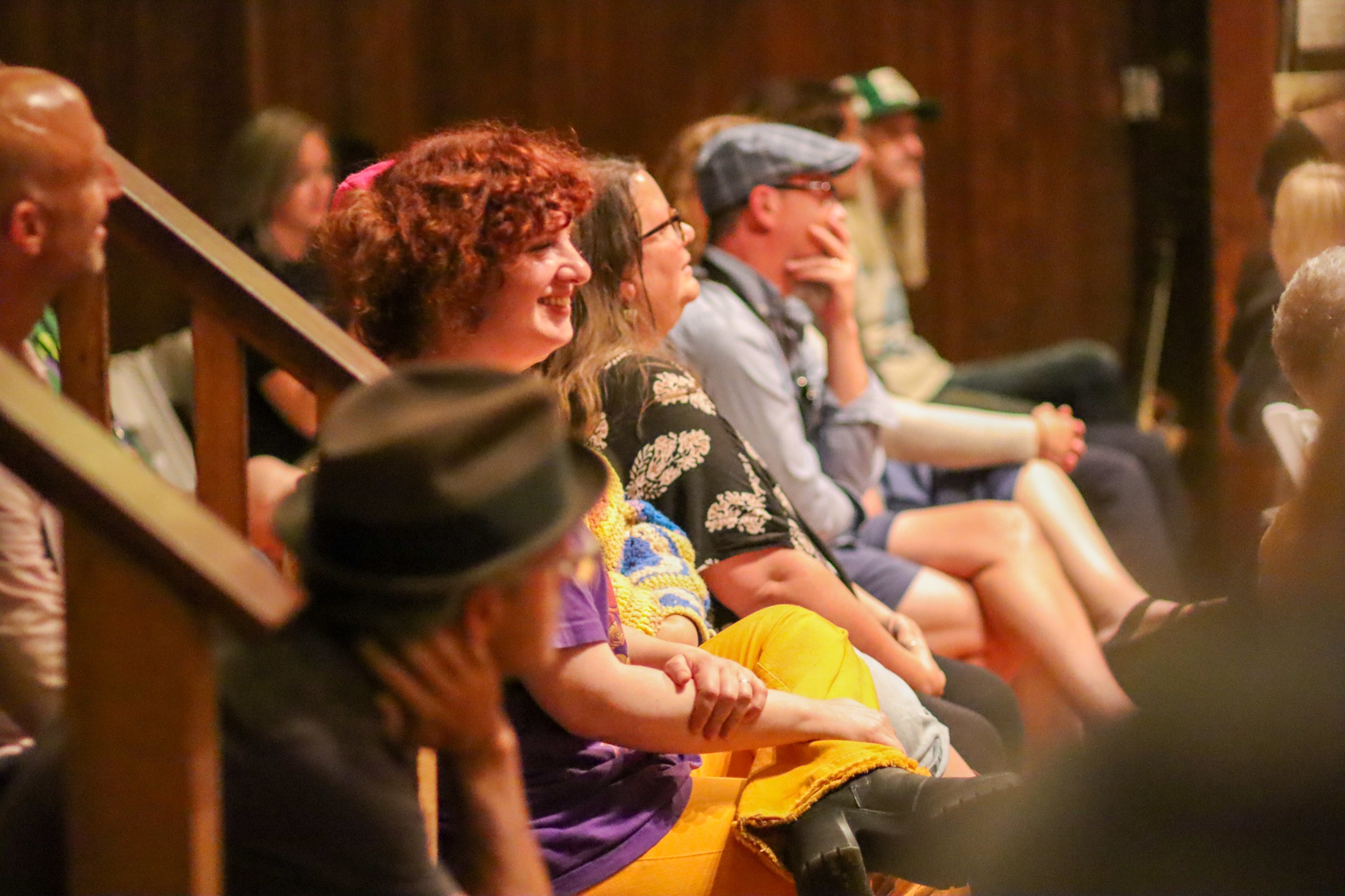Audience at an indoor event sitting on wooden benches, casually dressed, smiling and attentive.