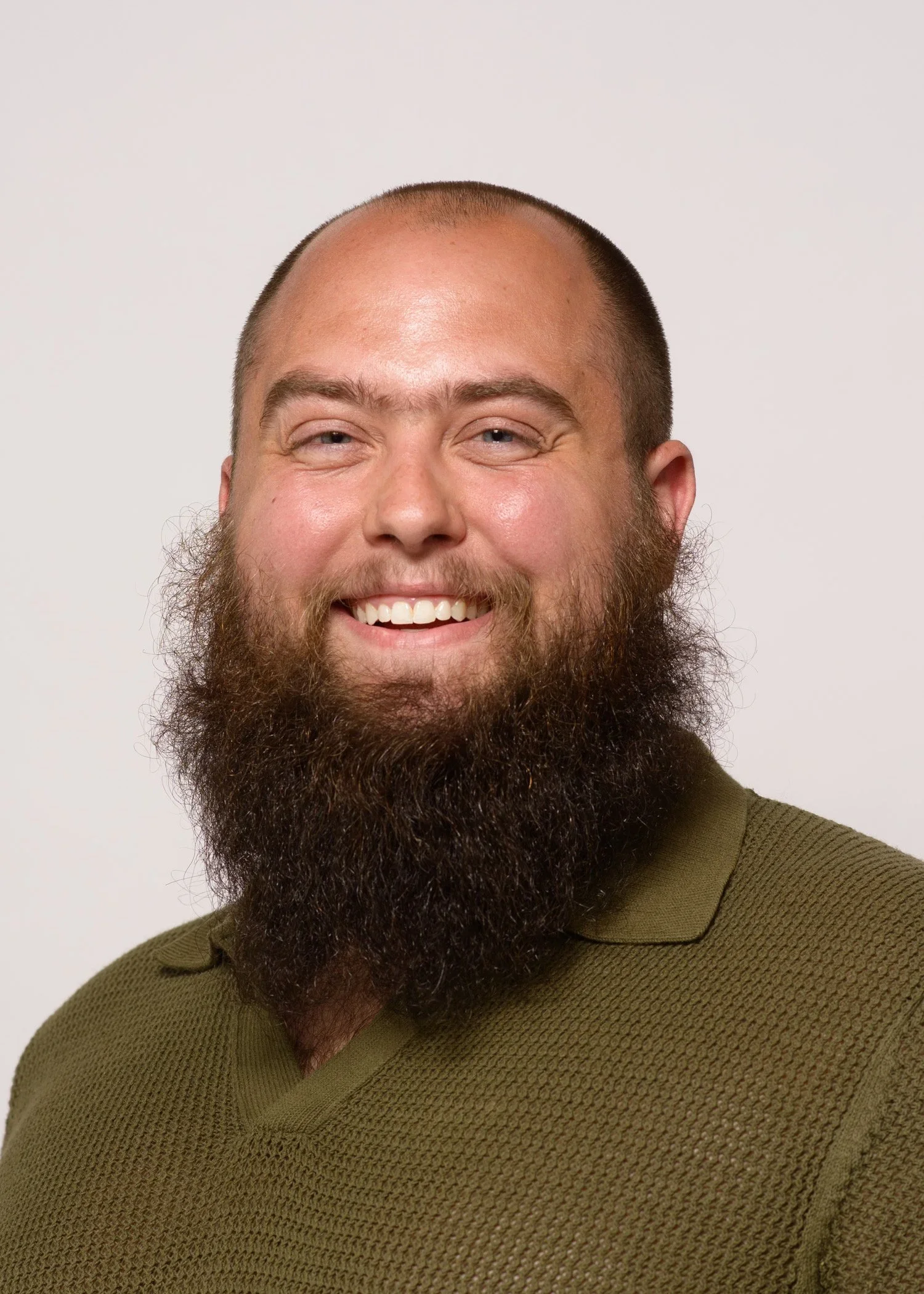 Close-up of a smiling man with a beard, wearing an olive green sweater, against a plain white background.
