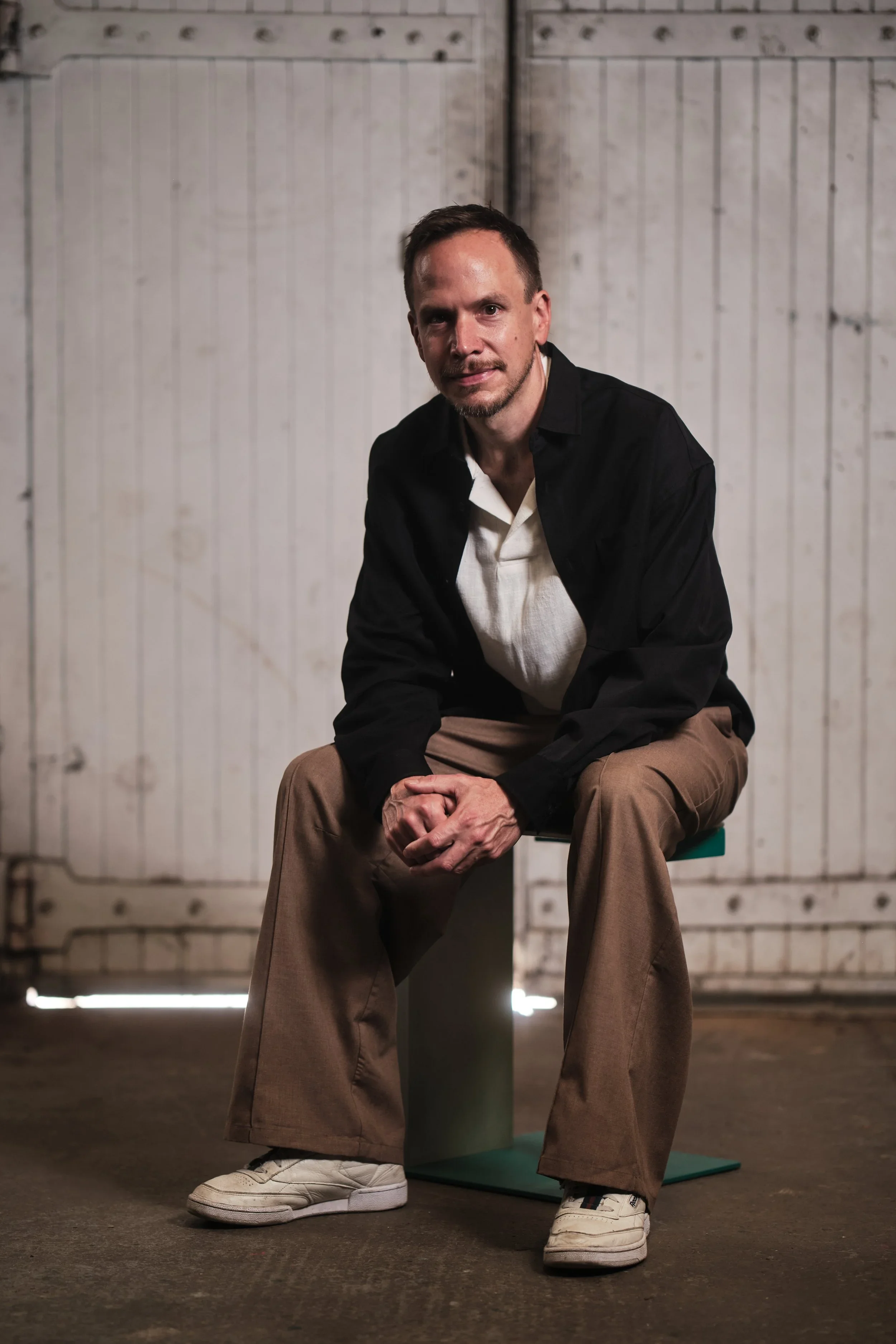 A man with short brown hair and a beard sitting on a stool in front of a whitewashed wooden wall. He is wearing a black jacket over a white shirt, beige pants, and white sneakers. The man is looking at the camera with a slight smile.