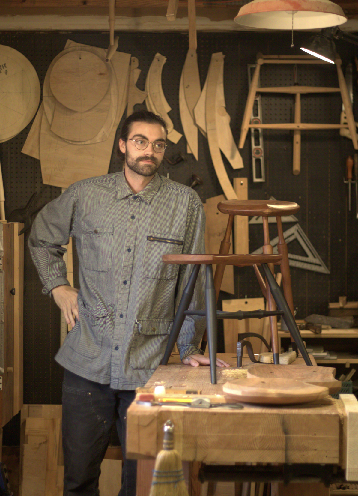 Man in a woodshop with woodworking tools and finished furniture, standing beside a wooden stool on a workbench.