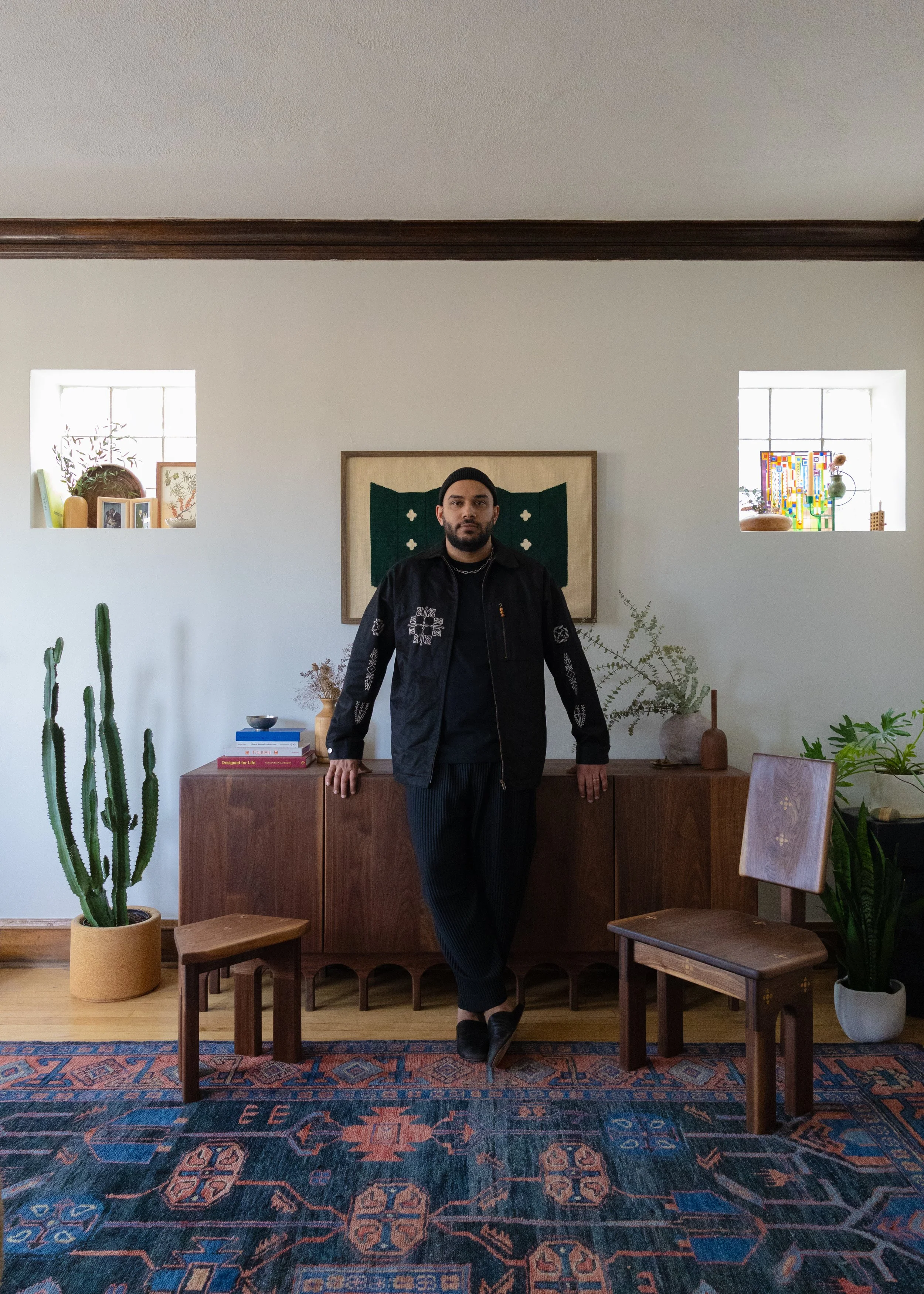 A man with a beard and black beanie wearing a black jacket with white embroidery, black shirt, and black pants, standing in a room with a white wall, wooden furniture, plants, and decorative items.