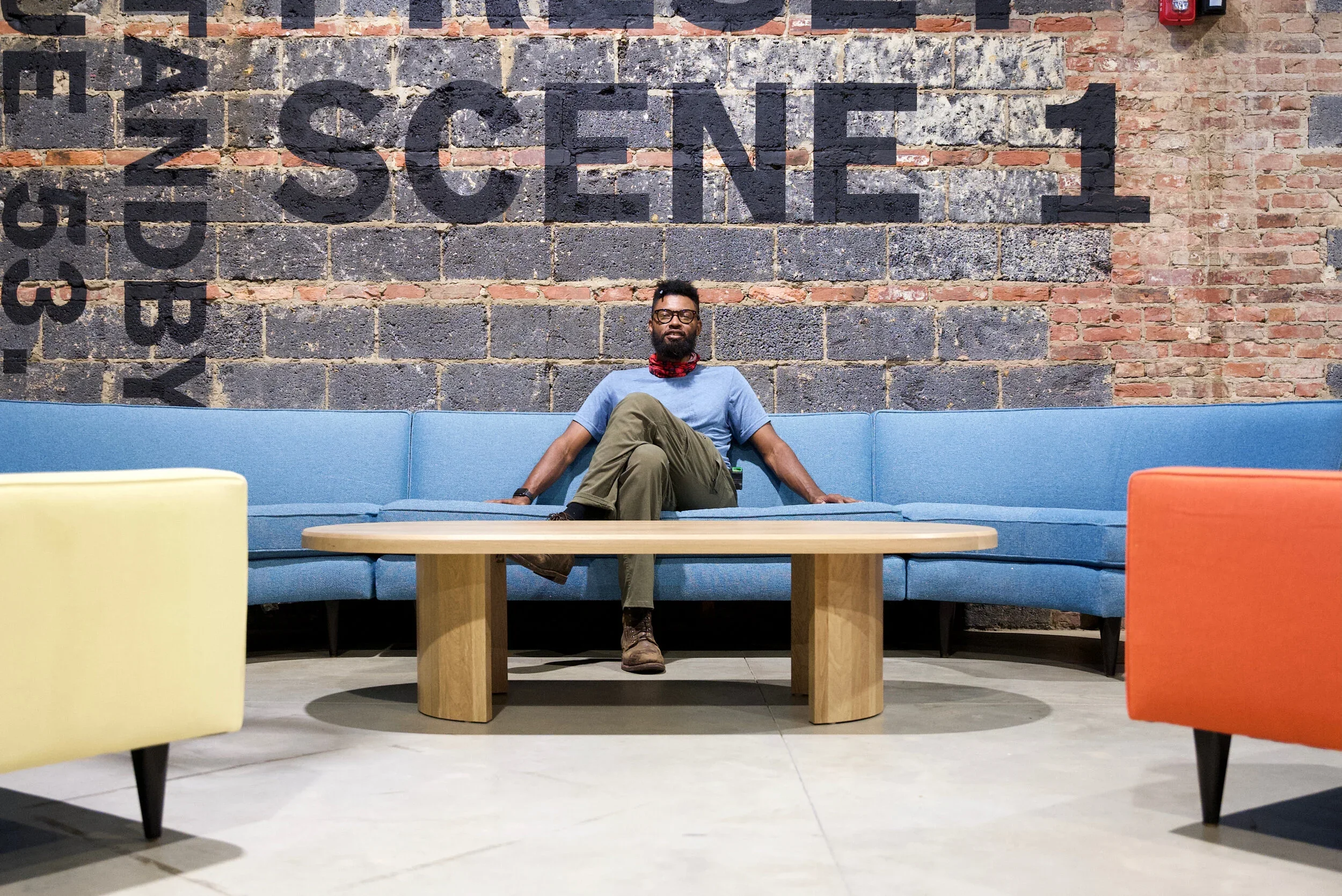 A man with a beard, glasses, and a red neck gaiter sits comfortably on a long blue cushioned bench in a modern indoor space, with a brick wall featuring large black painted letters reading 'SOCIETY' behind him. He is wearing a light blue t-shirt, khaki cargo pants, and brown boots, with his legs crossed and one arm resting on the bench.