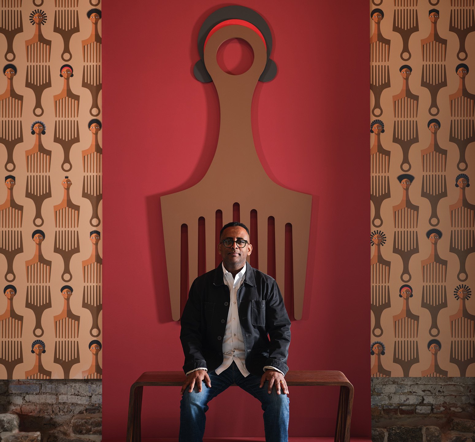 A man with glasses wearing a black jacket sitting on a wooden bench in front of a pink wall with a large abstract art piece resembling a fork with headphones. Two patterned wallpaper sections with stylized human faces in earthy tones flank the wall.