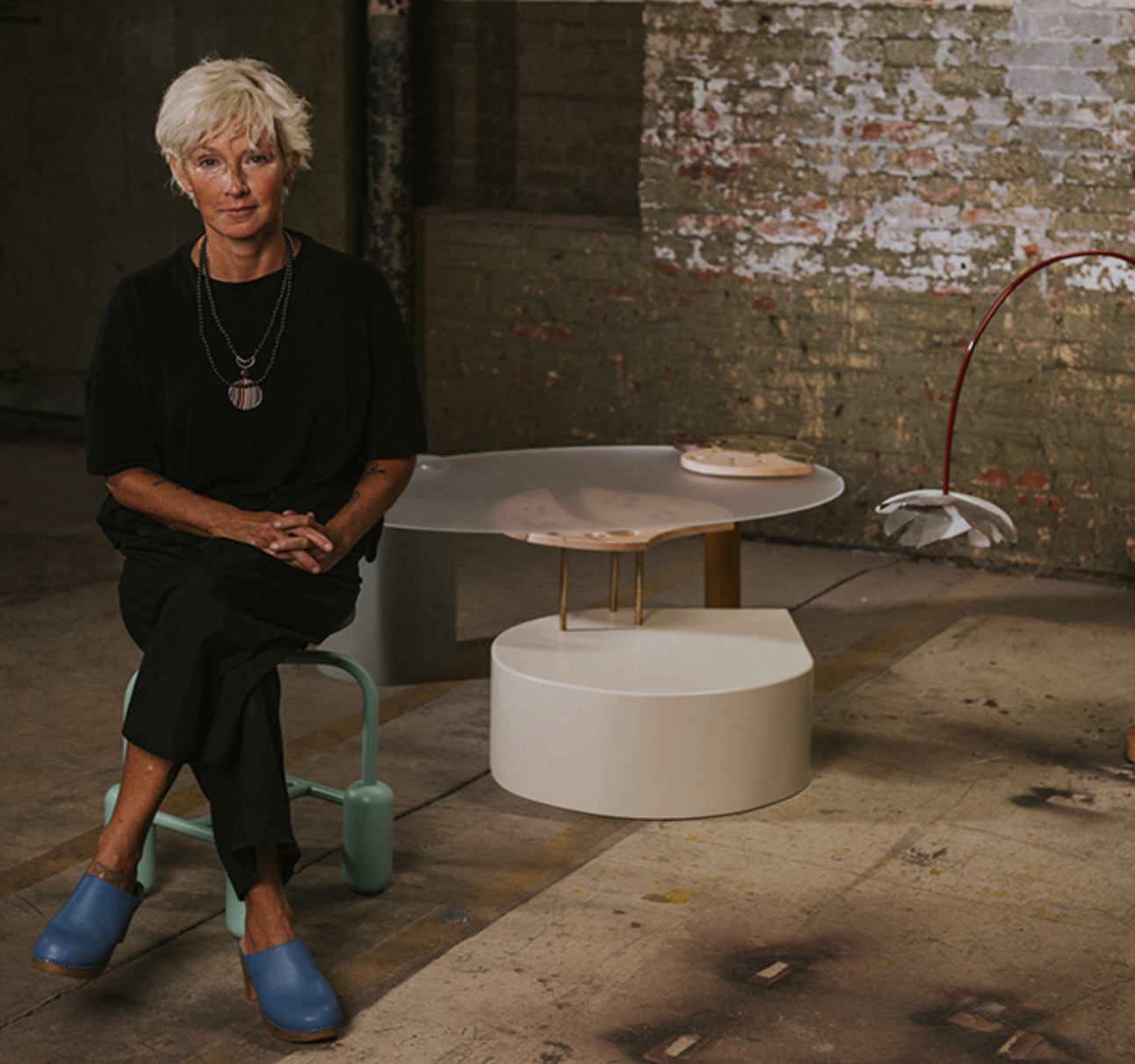 An older woman with short, white hair sitting on a modern teal stool in an industrial-style room with exposed brick walls. She is wearing a black outfit with layered necklaces and blue clogs. Behind her, there are contemporary tables and a unique floor lamp shaped like a flower.