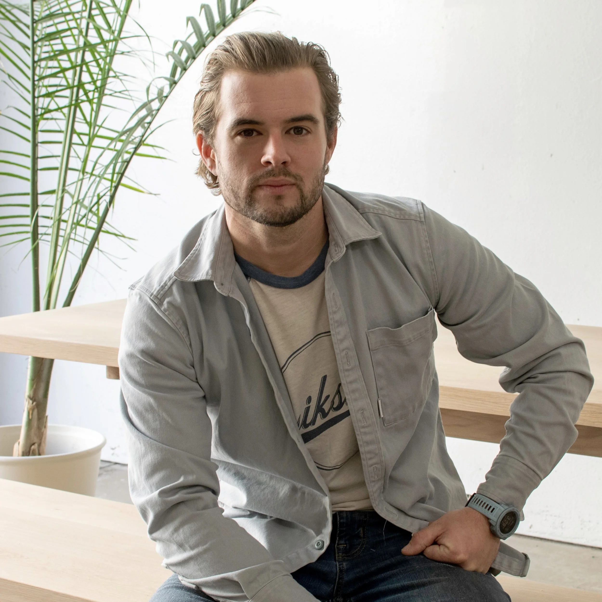 A man with brown hair and a beard, wearing a light gray jacket and dark jeans, sitting at a desk with a potted plant in the background.