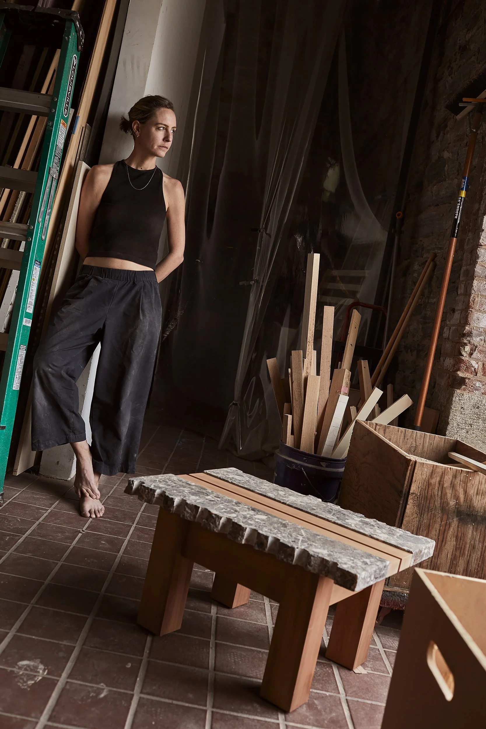 A woman with short hair and a black sleeveless top stands barefoot in a workshop, leaning against the wall. The workshop contains wooden boards, a ladder, tools, and large containers of wood pieces, with brick walls and tiled flooring.