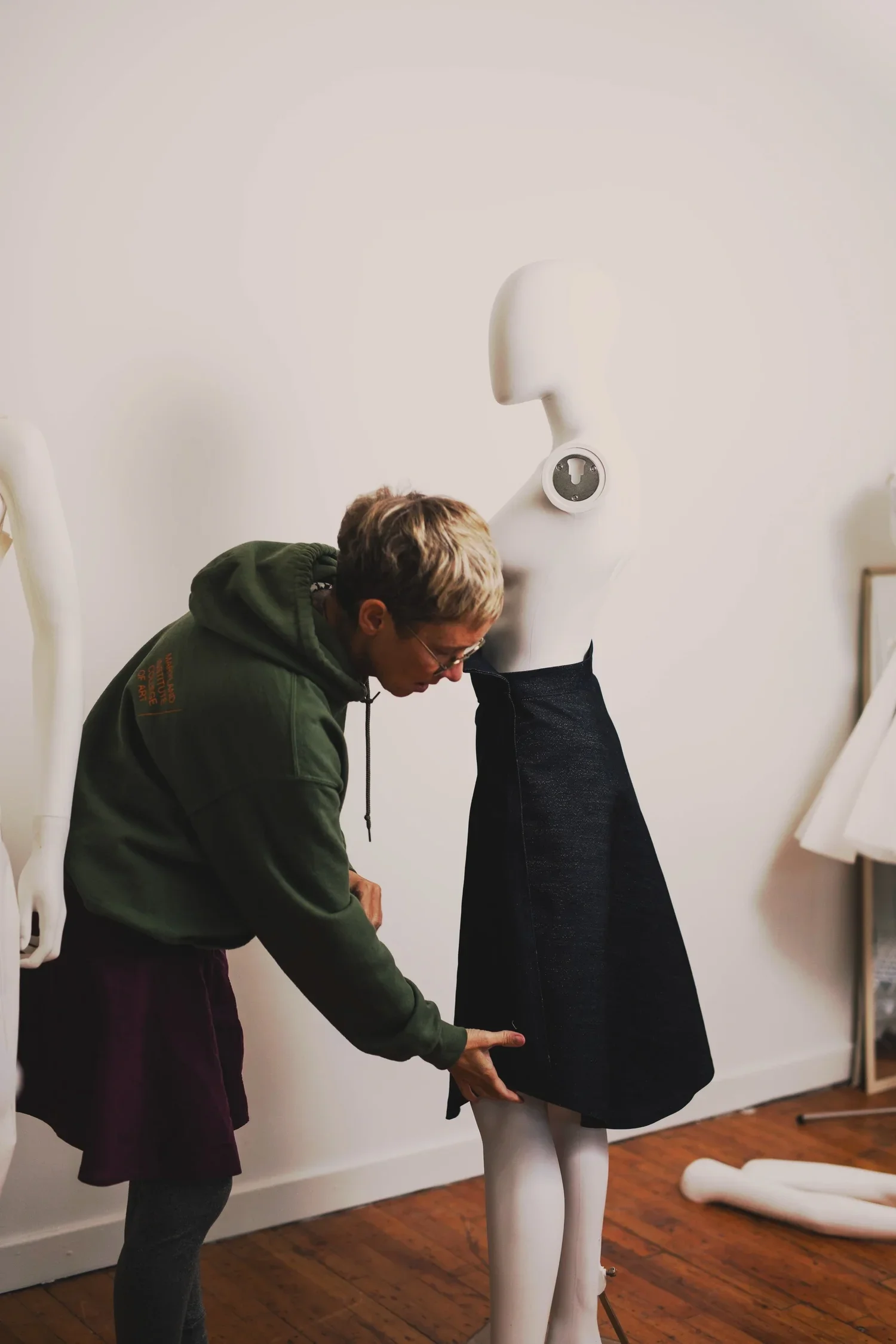 A person with short blond hair wearing glasses and a green hoodie is leaning over and pointing at a black dress on a white mannequin in a room with wooden floors and white walls.