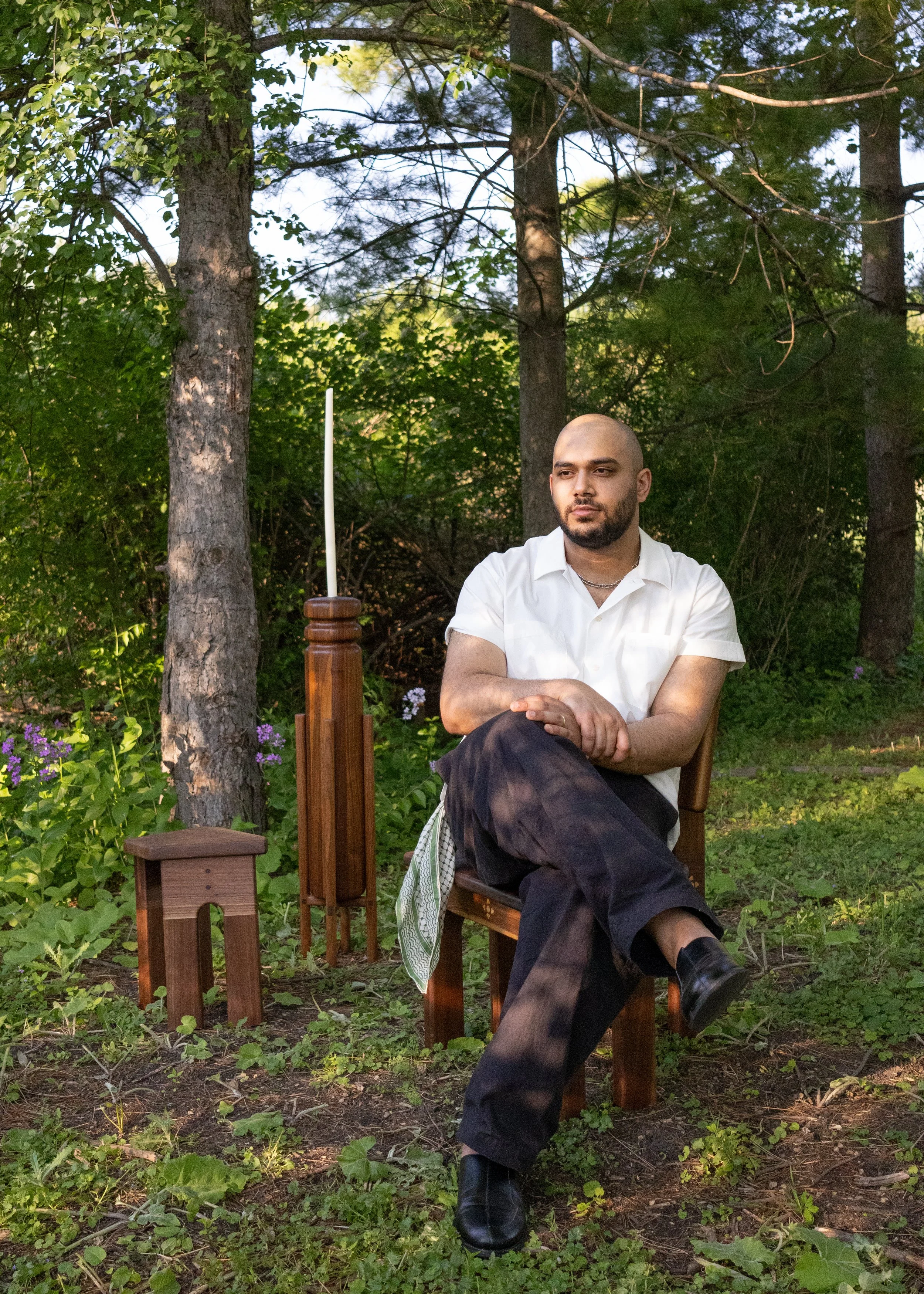 A man with a shaved head, beard, wearing a white shirt and black pants, sitting outdoors in a wooded area with trees and green foliage. He is seated on a wooden chair with his arms crossed over his lap, with a calm expression. There are small purple flowers and green plants on the ground, and a white candlestick on a wooden stand nearby.