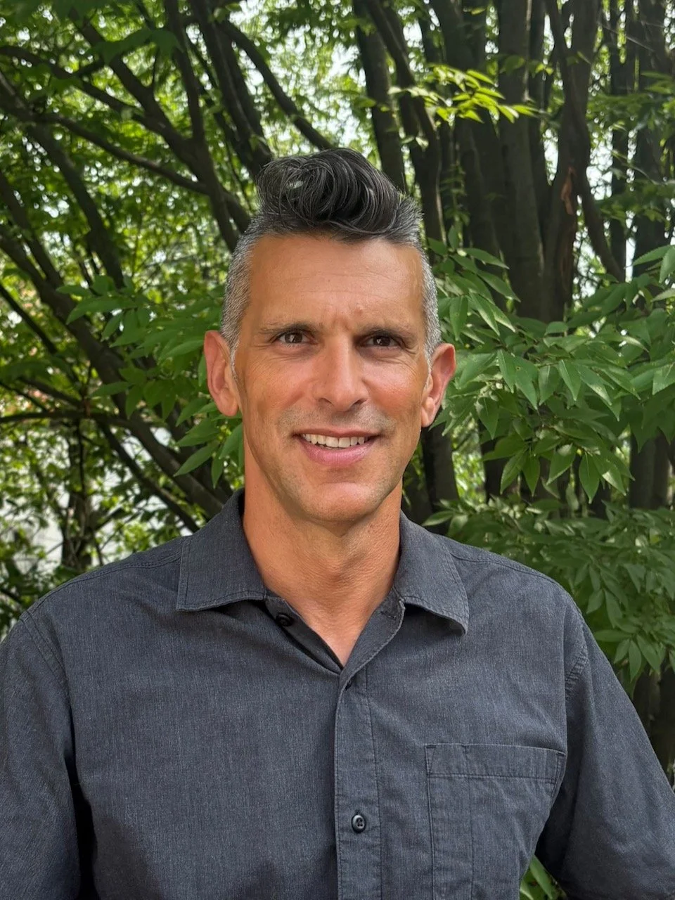 A man with short gray and black hair standing outdoors in front of green leafy trees, smiling at the camera, wearing a dark gray button-up shirt.