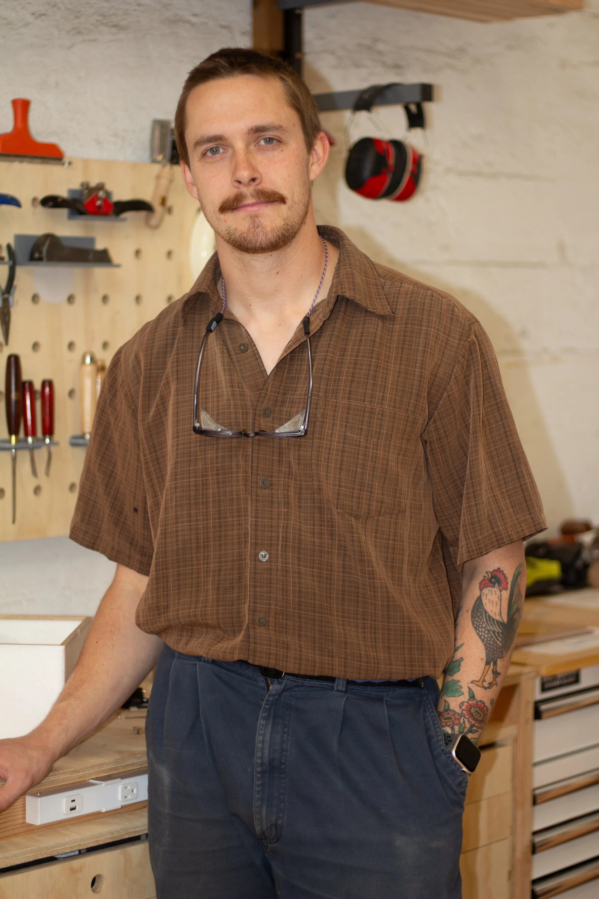A man with a mustache and tattoos, wearing a brown checkered shirt with sunglasses around his neck, standing in a workshop with tools on the wall behind him.
